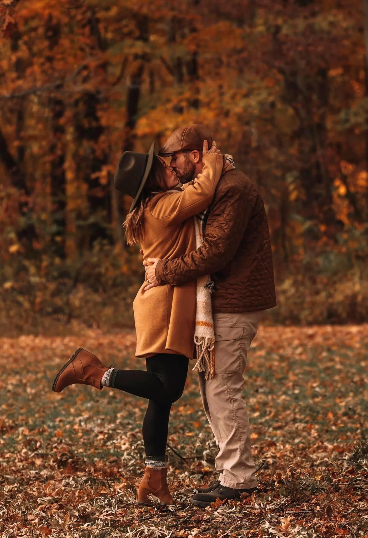 A couple kissing in a fall forest with autumn leaves on the ground and trees in the background.