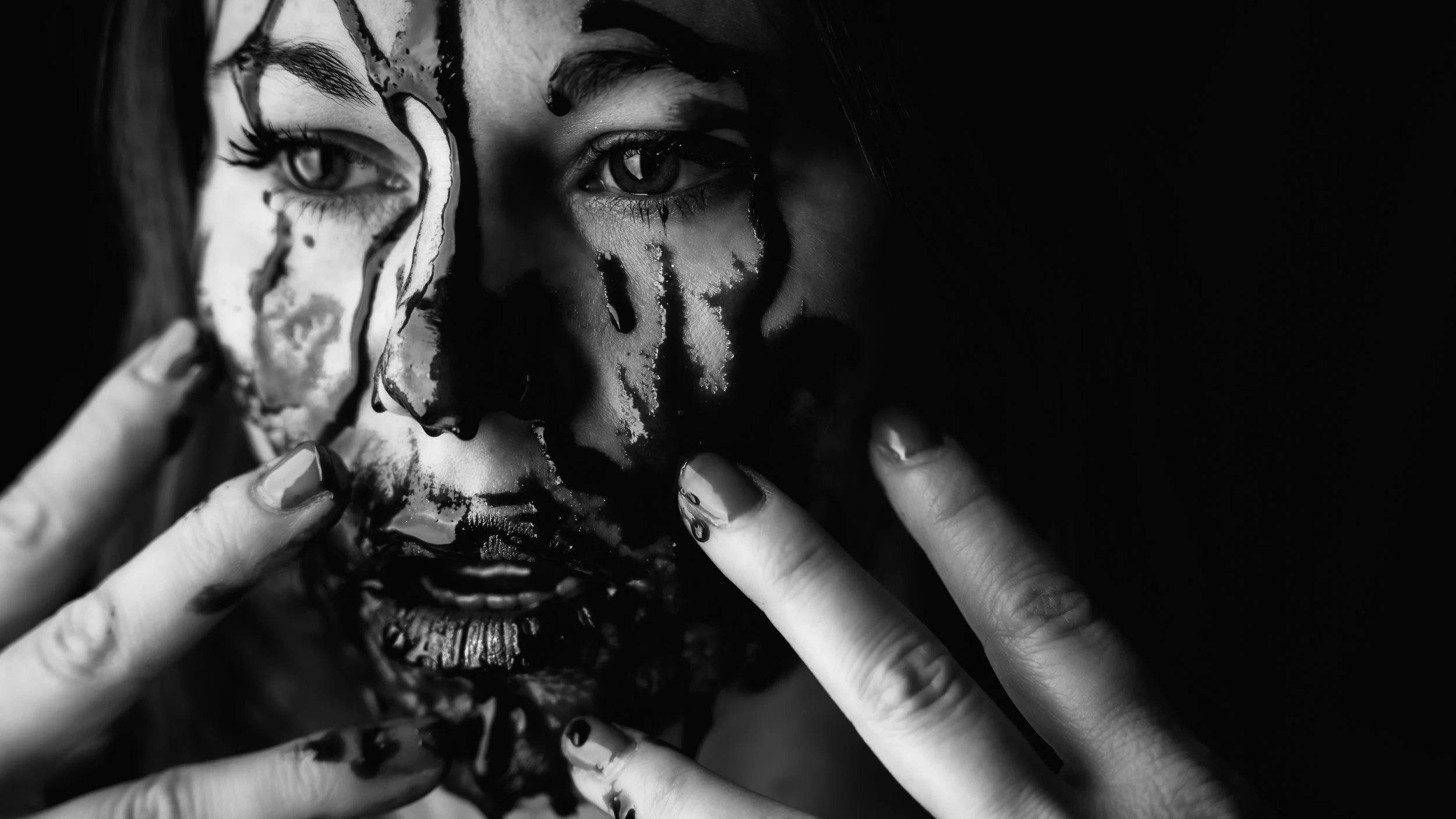 A woman with dripping paint on her face, holding her fingers near her lips, in black and white.