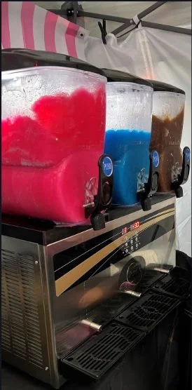 Three large beverage slurpee machines with colorful drinks—pink, blue, and brown—placed on a commercial machine, with a striped pink and white tent in the background.