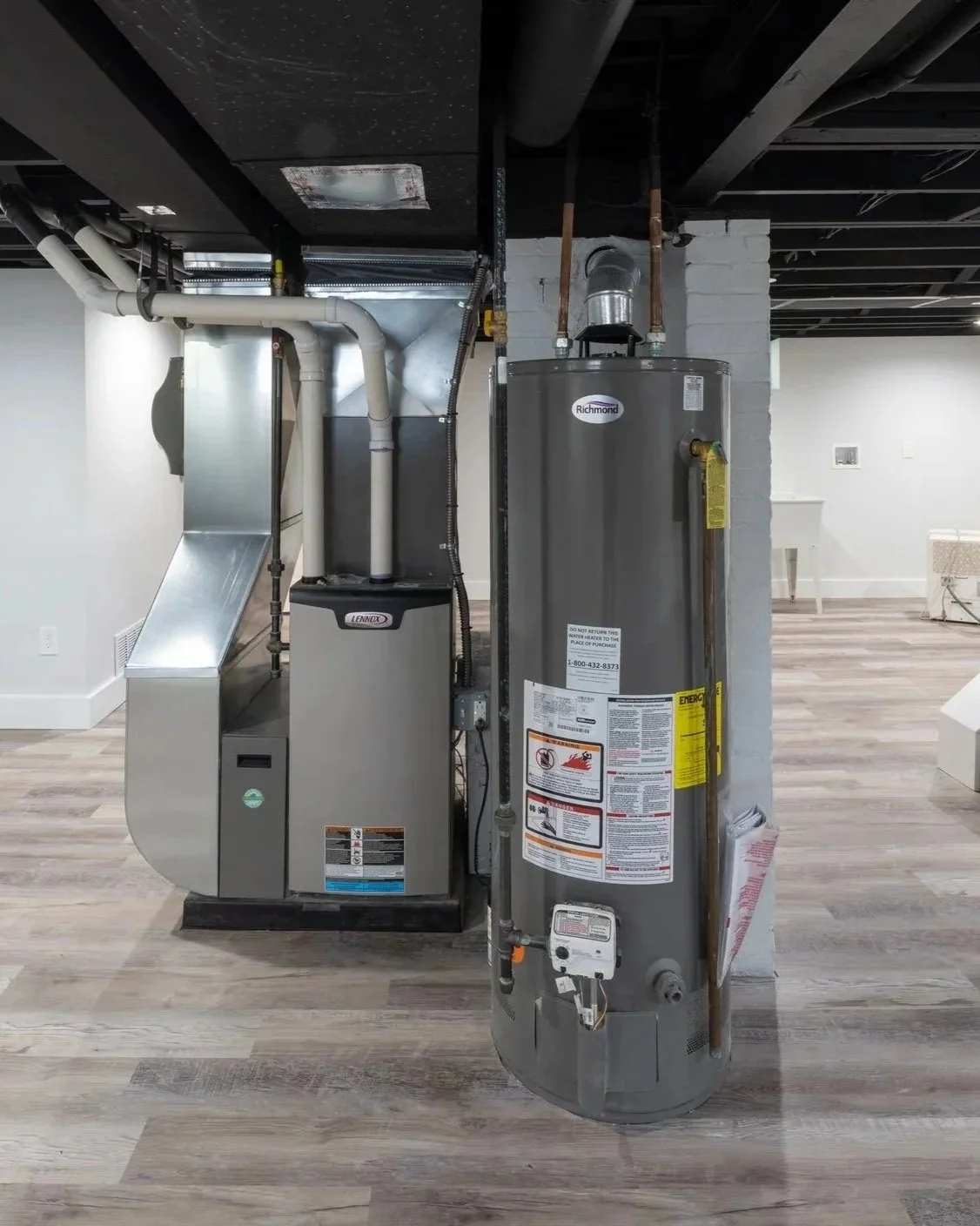 Water heater and furnace in a basement with wooden flooring and white walls.