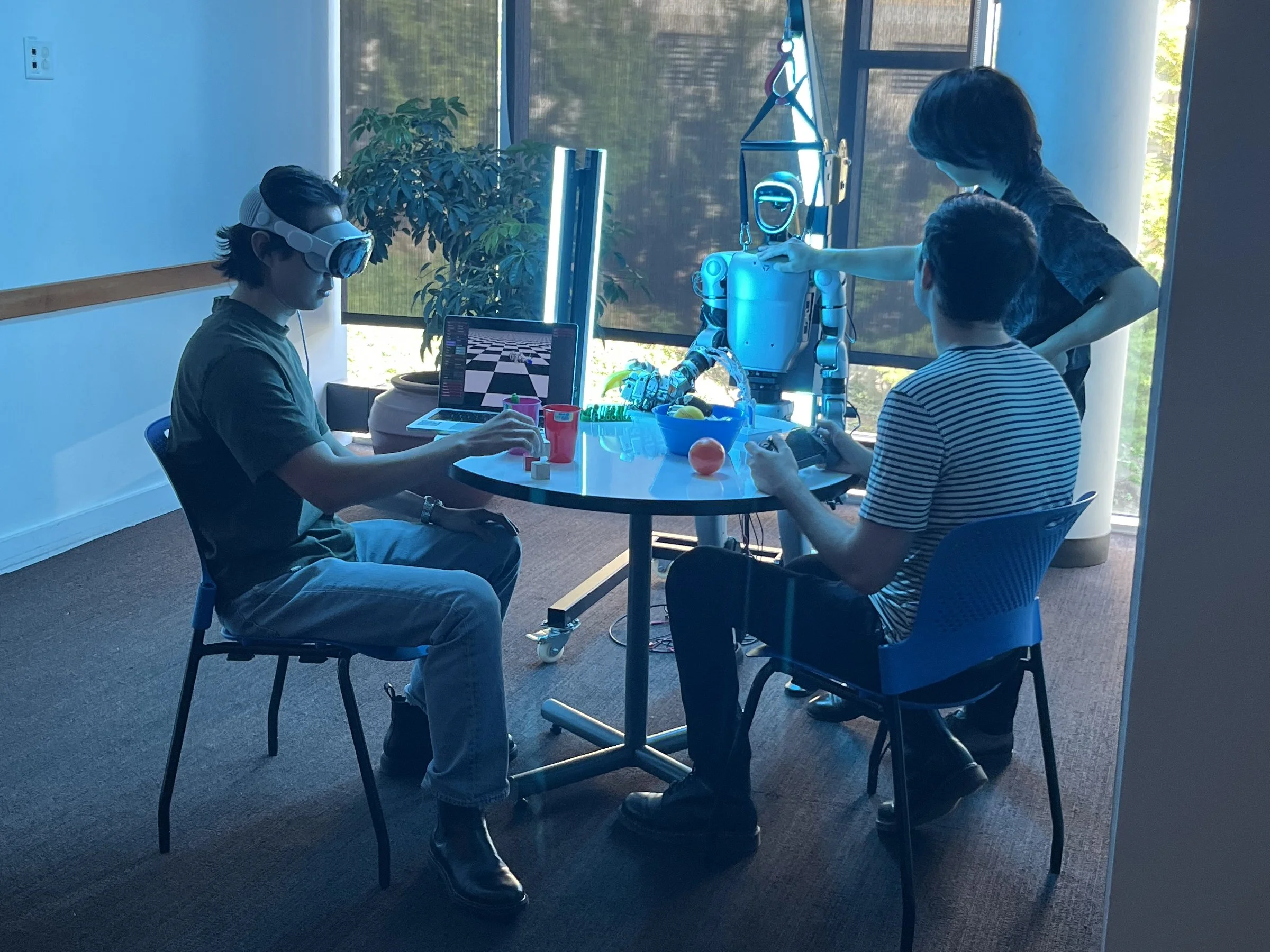 Three people in a room with large windows, playing with a humanoid robot while one person wearing virtual reality goggles observes, and a laptop with a checkered game on the screen is on the table along with some cups and fruit.