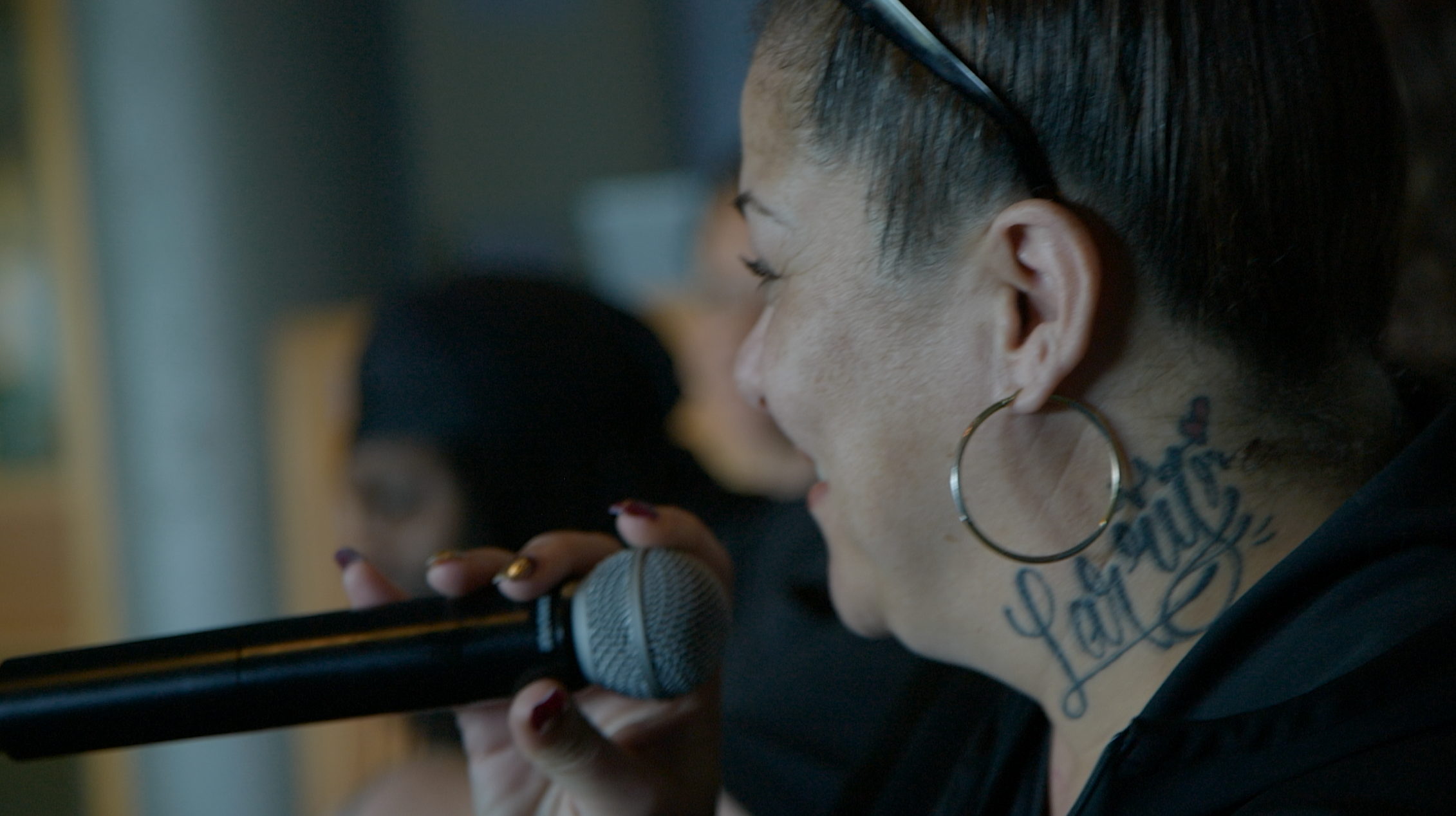 A woman with glasses, hoop earrings, and tattoos on her neck, singing into a microphone with eyes closed, in an indoor setting.