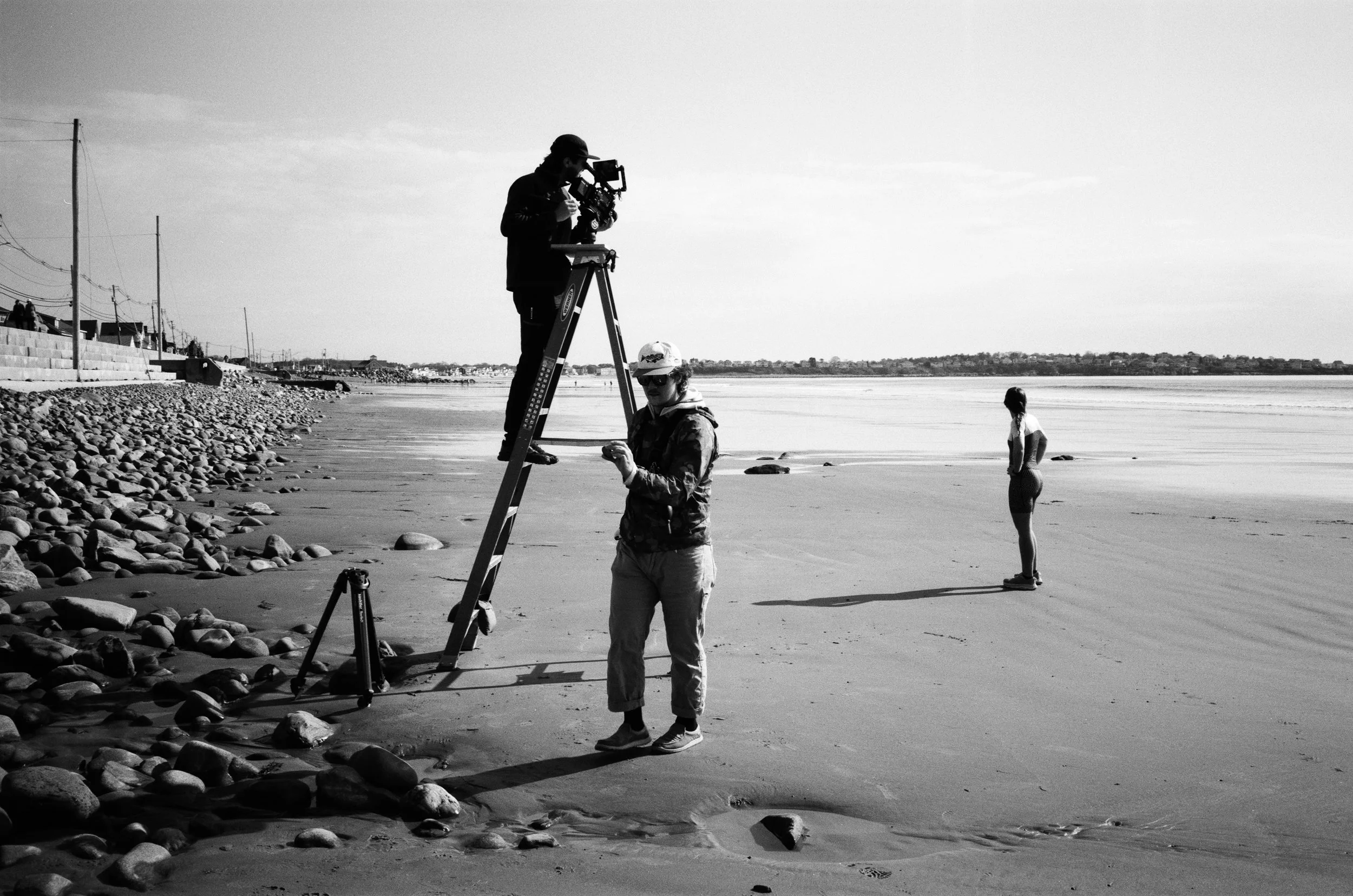 Group of people filming at the beach with a camera on a tripod, one person standing on a ladder, others on the ground, in a black-and-white photo.