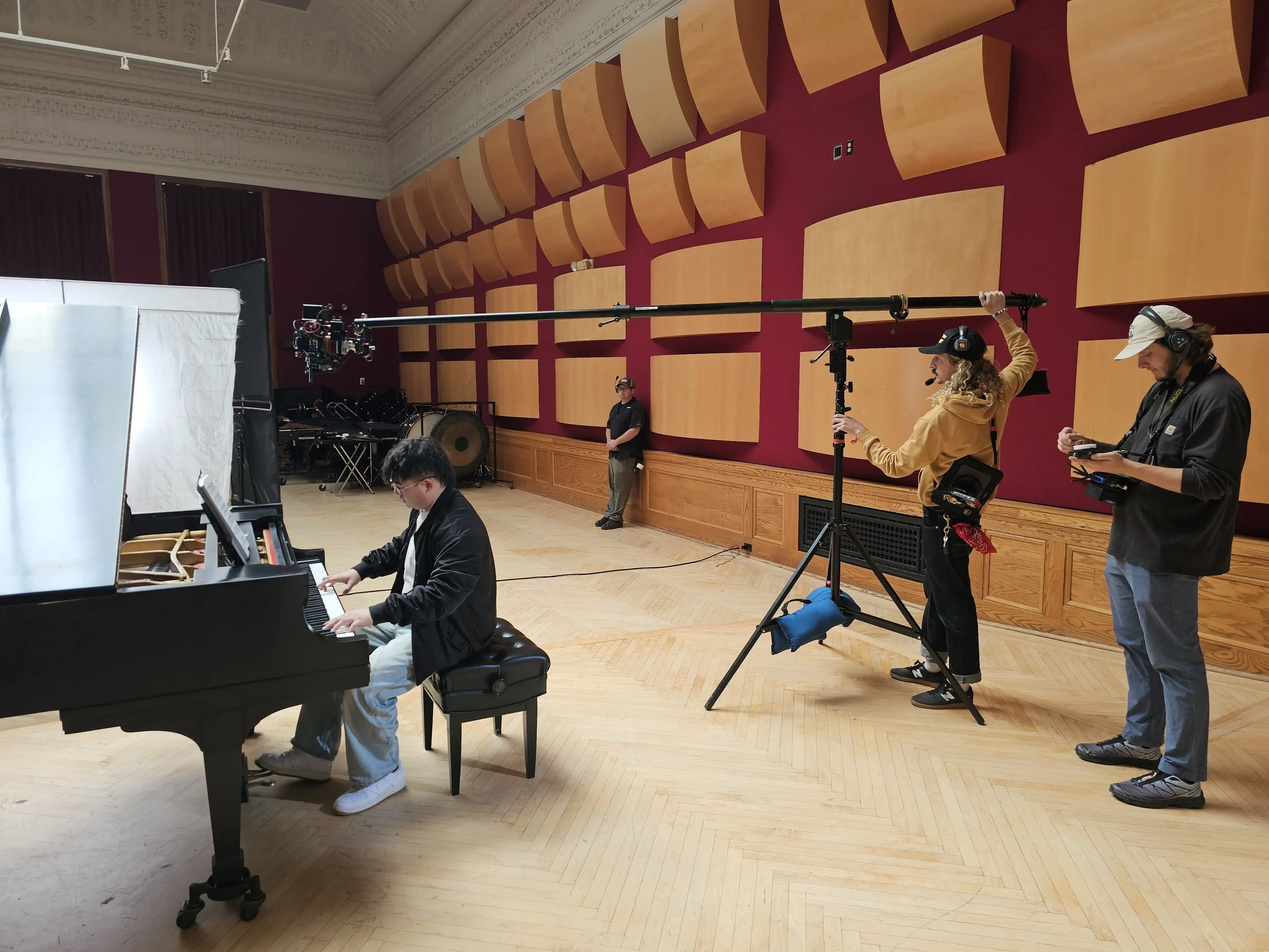 Young man playing grand piano in a recording studio with soundproofing panels, camera crew filming, and crew members observing.