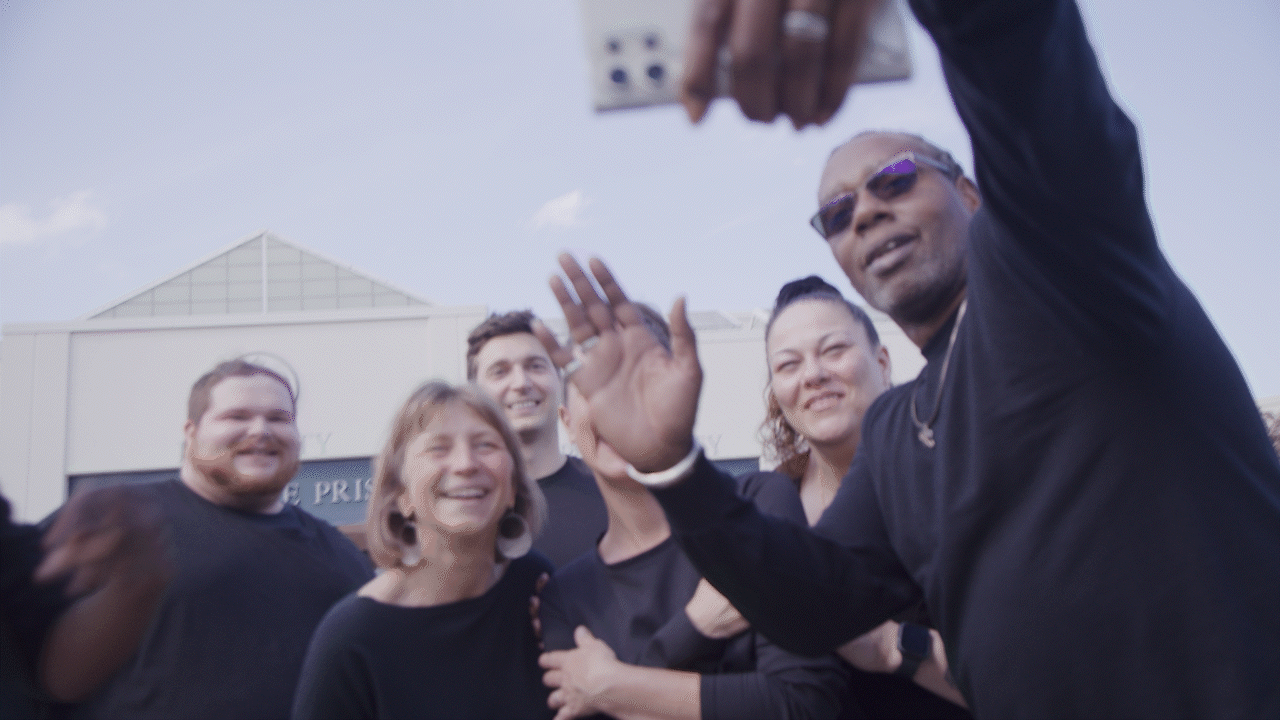 A group of six diverse people smiling and waving at the camera during an outdoor event on a sunny day.