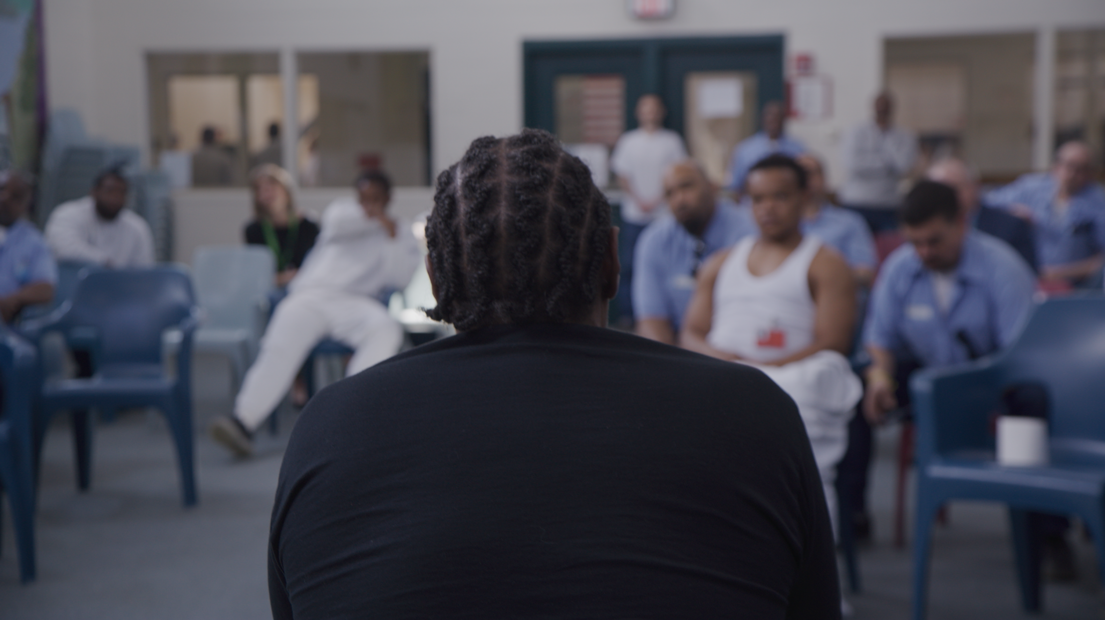 Person with braided hair sitting in front of a group of people in a classroom or meeting space.