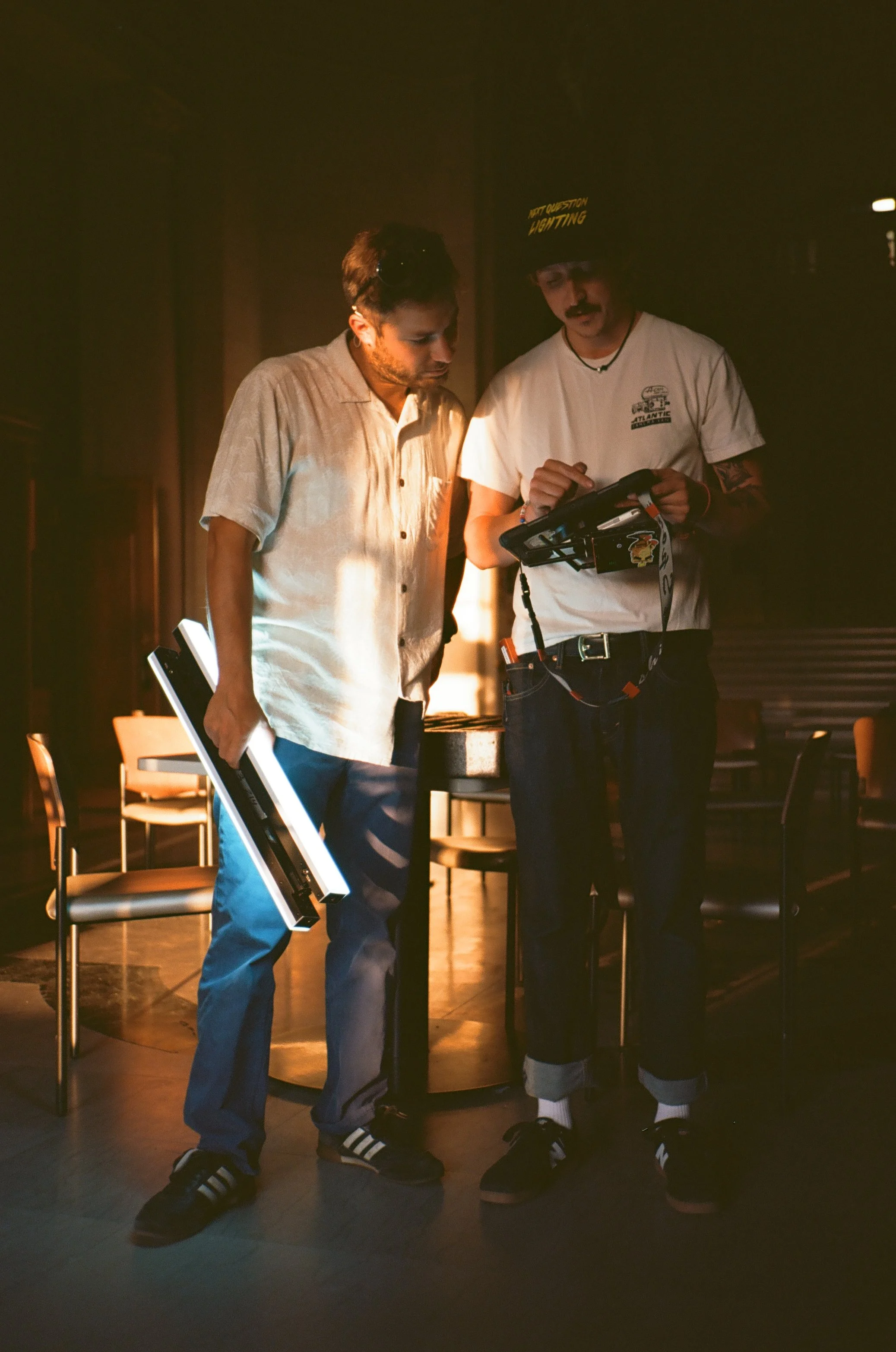 Two young men are standing close to each other in a dimly lit room, looking at a tablet device held by the man on the right. The man on the left has dark hair and is wearing a beige short-sleeved shirt and blue jeans. The man on the right has a mustache, tattoos on his arms, and is wearing a white t-shirt, dark jeans, a black wide-brimmed hat with yellow text, and a necklace. There are chairs and a table in the background.