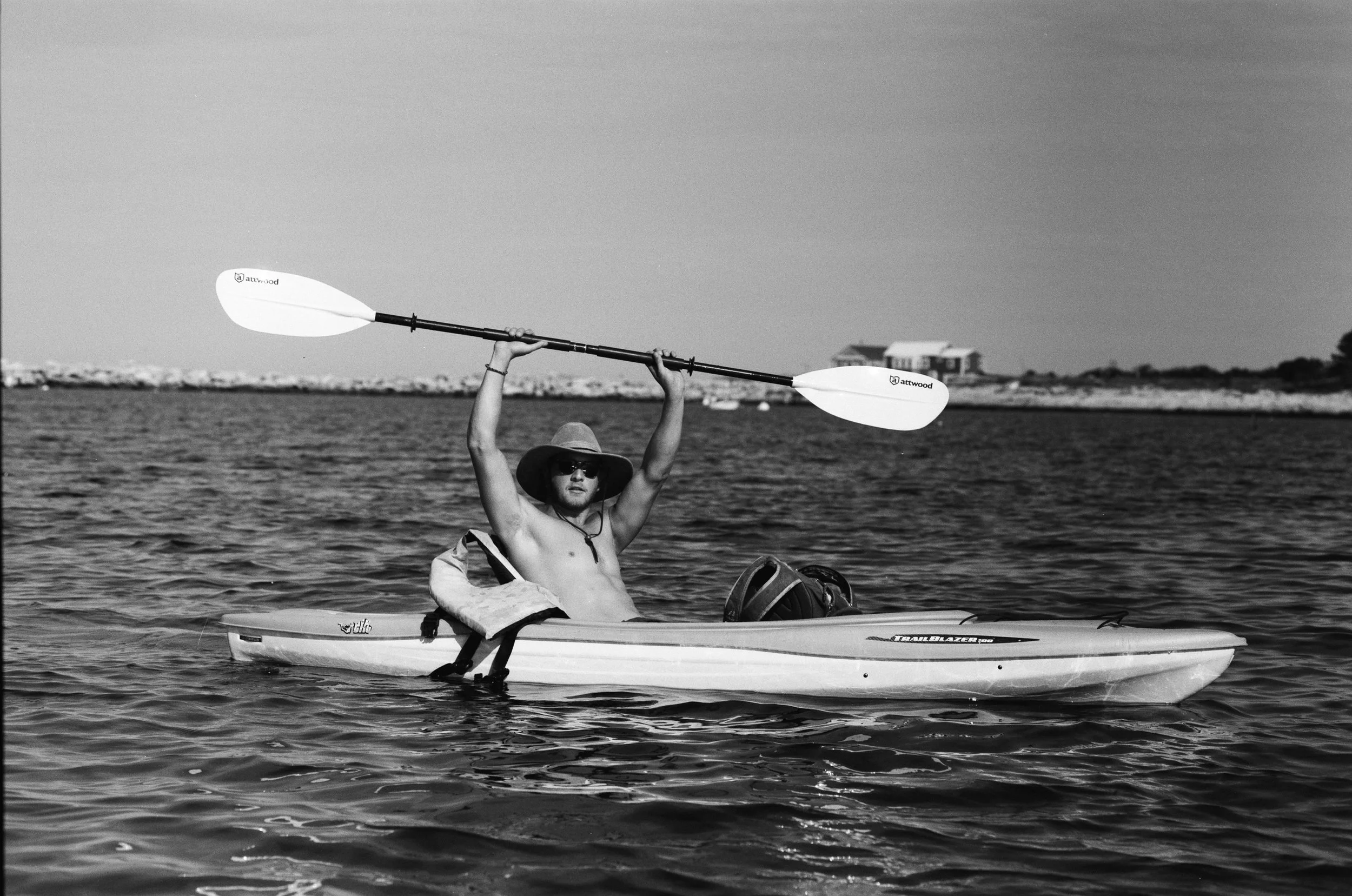 A shirtless man wearing sunglasses and a hat sitting in a kayak, holding a double-ended paddle above his head, on a body of water with a background of boats and buildings.