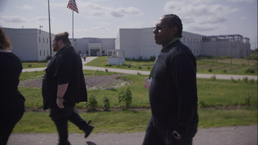 People walking outdoors near a white building, with an American flag flying in the background.