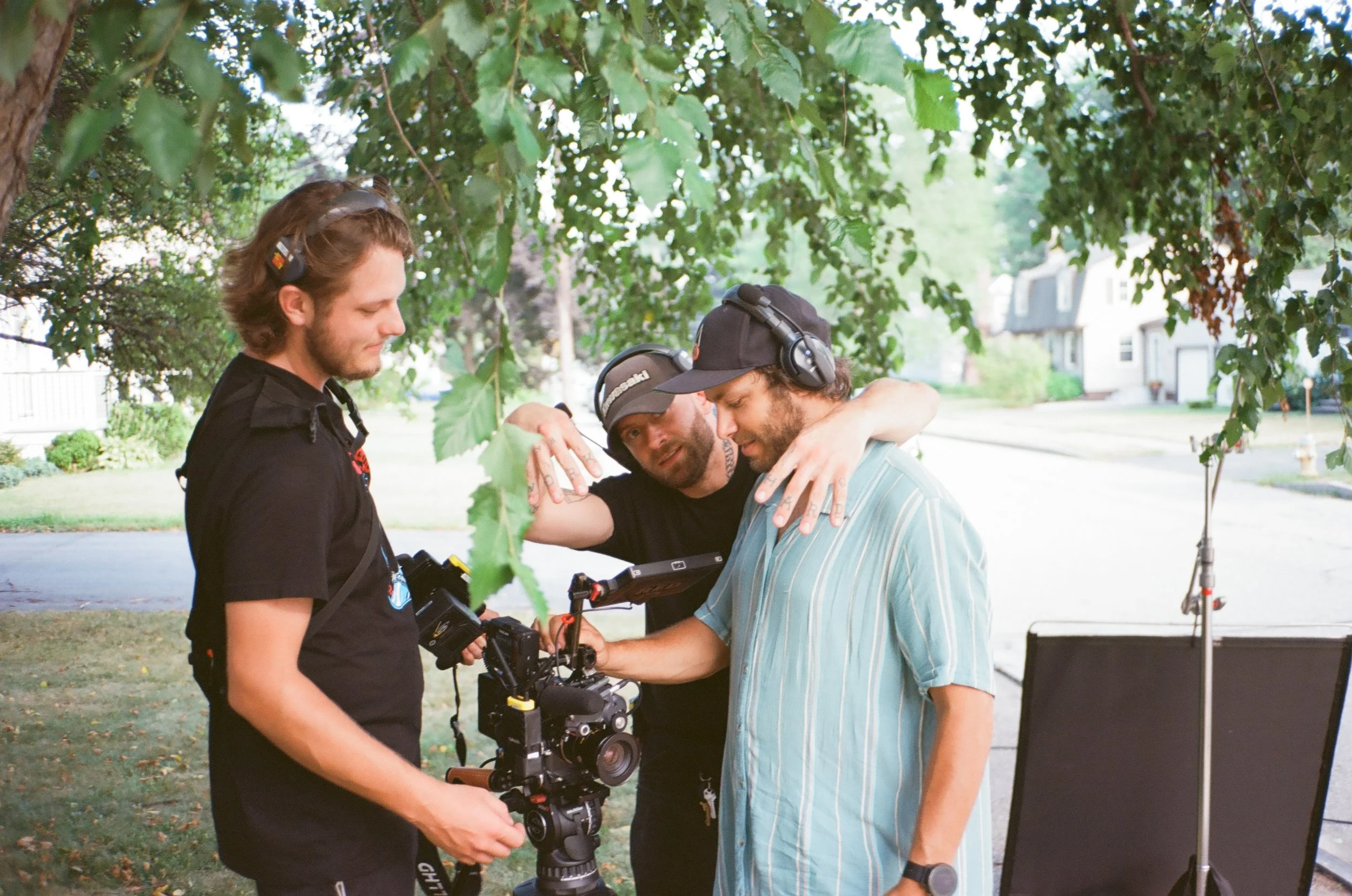 Three men are working together outdoors, looking at a camera mounted on a stabilizer. They appear focused on their task, under a tree with houses in the background.