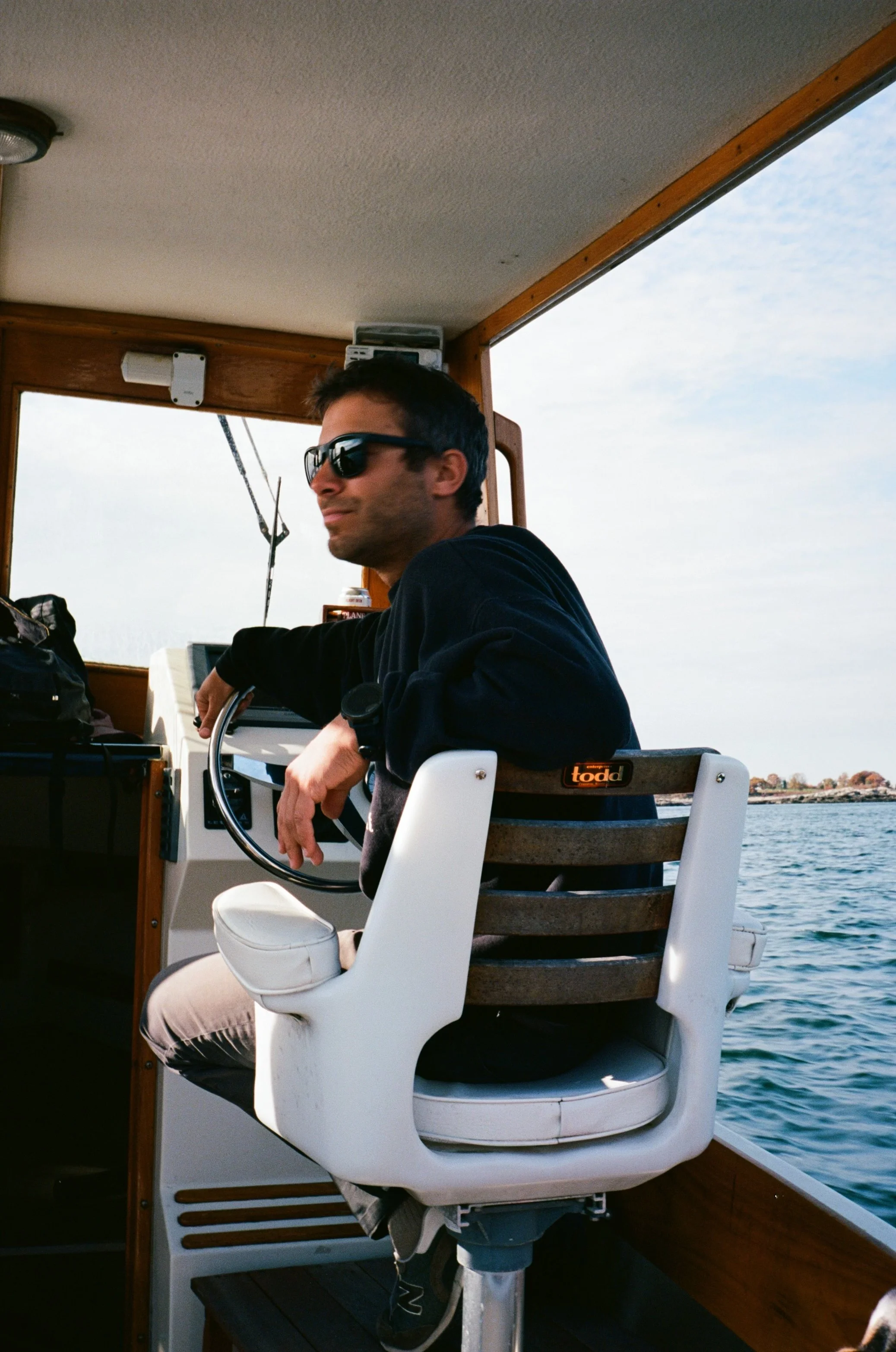 A man with dark hair, wearing sunglasses, a black shirt, beige pants, and black shoes, sitting on a white boat seat. He is holding onto a ship's steering wheel with his left hand. The boat is on a body of water, and there are buildings in the distant background. The inside of the boat has wooden trim and various equipment.