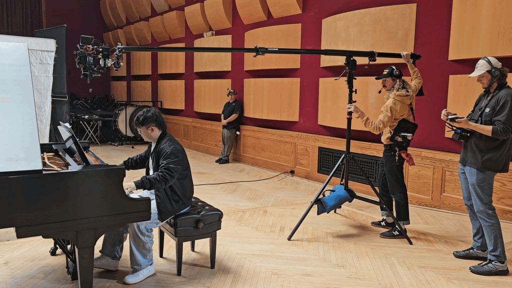 A film crew is filming a scene in a music recording studio with a pianist playing a grand piano. The crew includes a cameraman with a monitor, a person holding a boom microphone, and another crew member with a remote control. A person is standing against the wall observing.