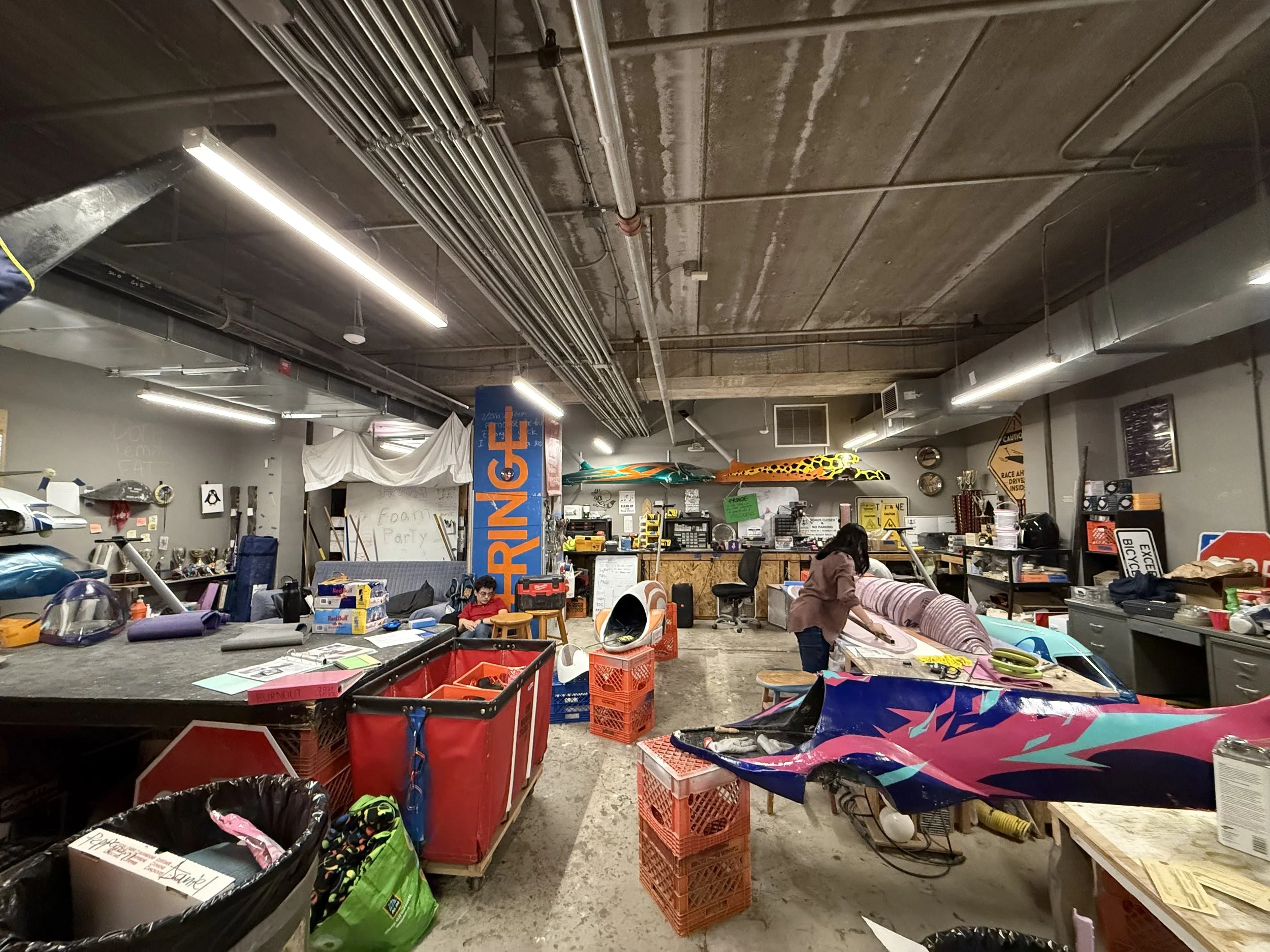A cluttered hobby workshop with various supplies and equipment. The ceiling reveals exposed pipes and concrete. There are people working at tables, and numerous colorful items including model planes, signs, and containers are scattered around.