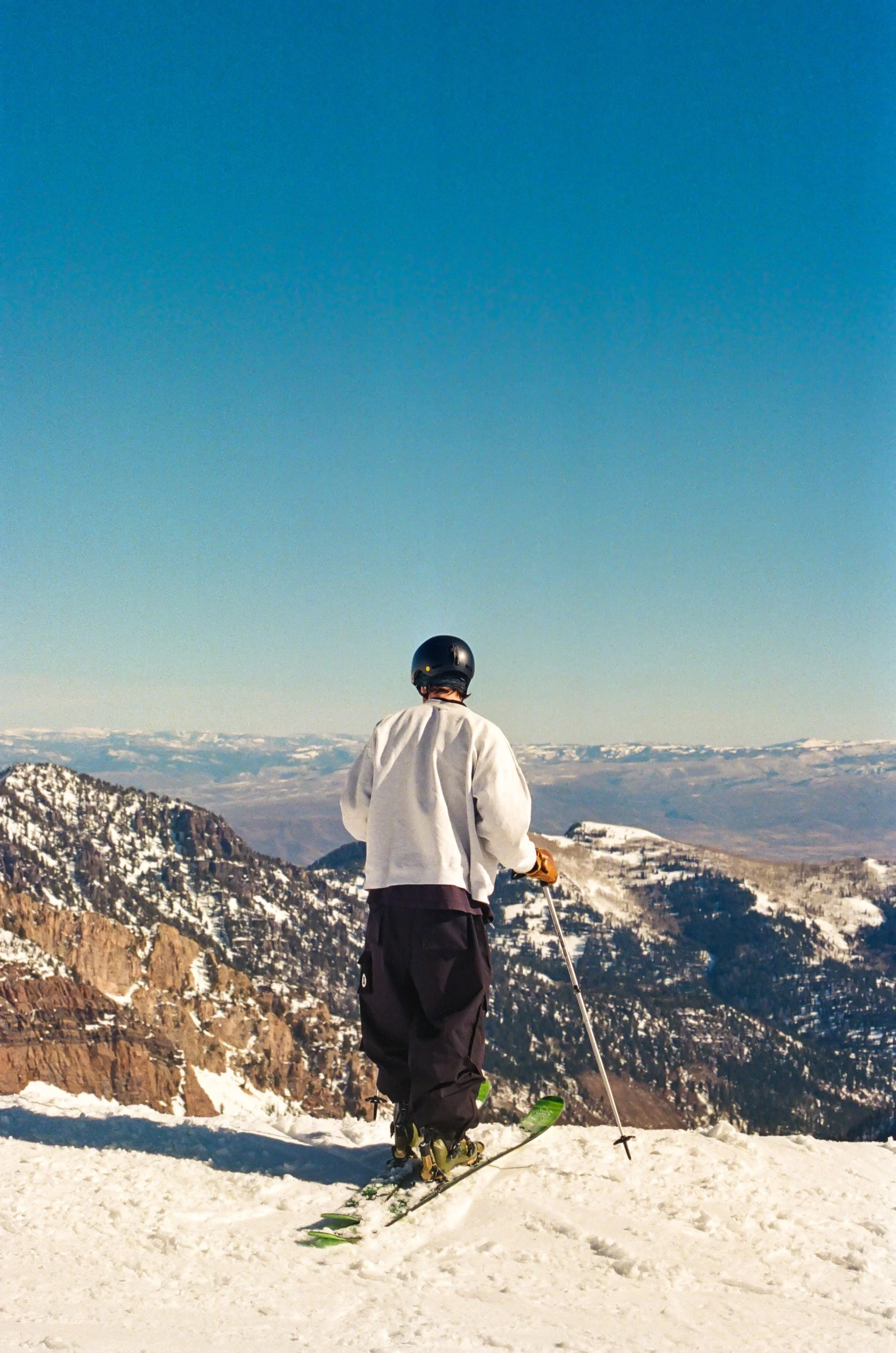 Person skiing on snow-covered mountain with rocky cliffs and distant mountain ranges under a blue sky.