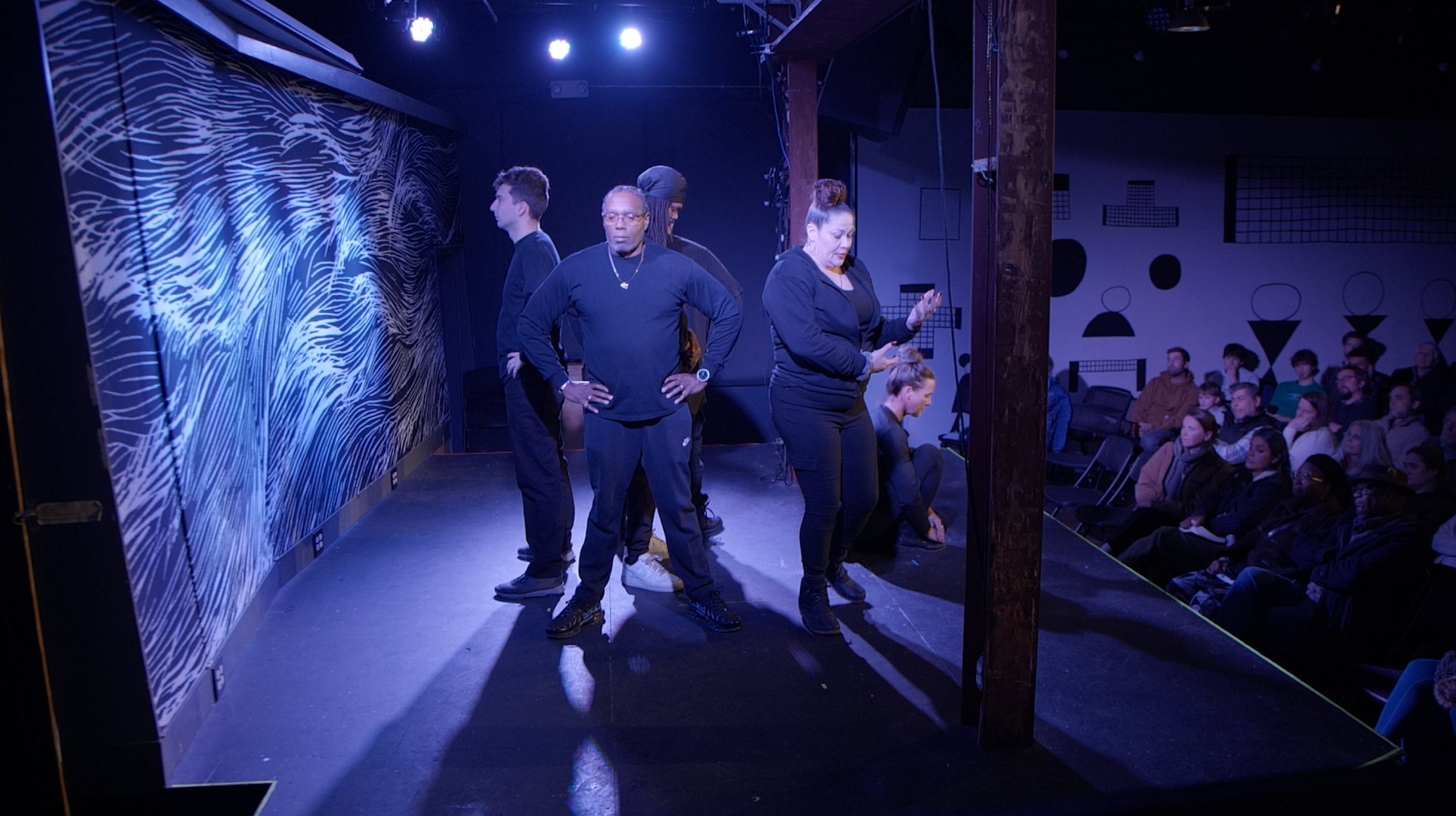 Four performers are standing on a stage, with three of them facing to the left and one looking down at her phone. The audience is seated in front of the stage, and the background features a wall with geometric and abstract designs. The lighting is dim with blue and purple hues.