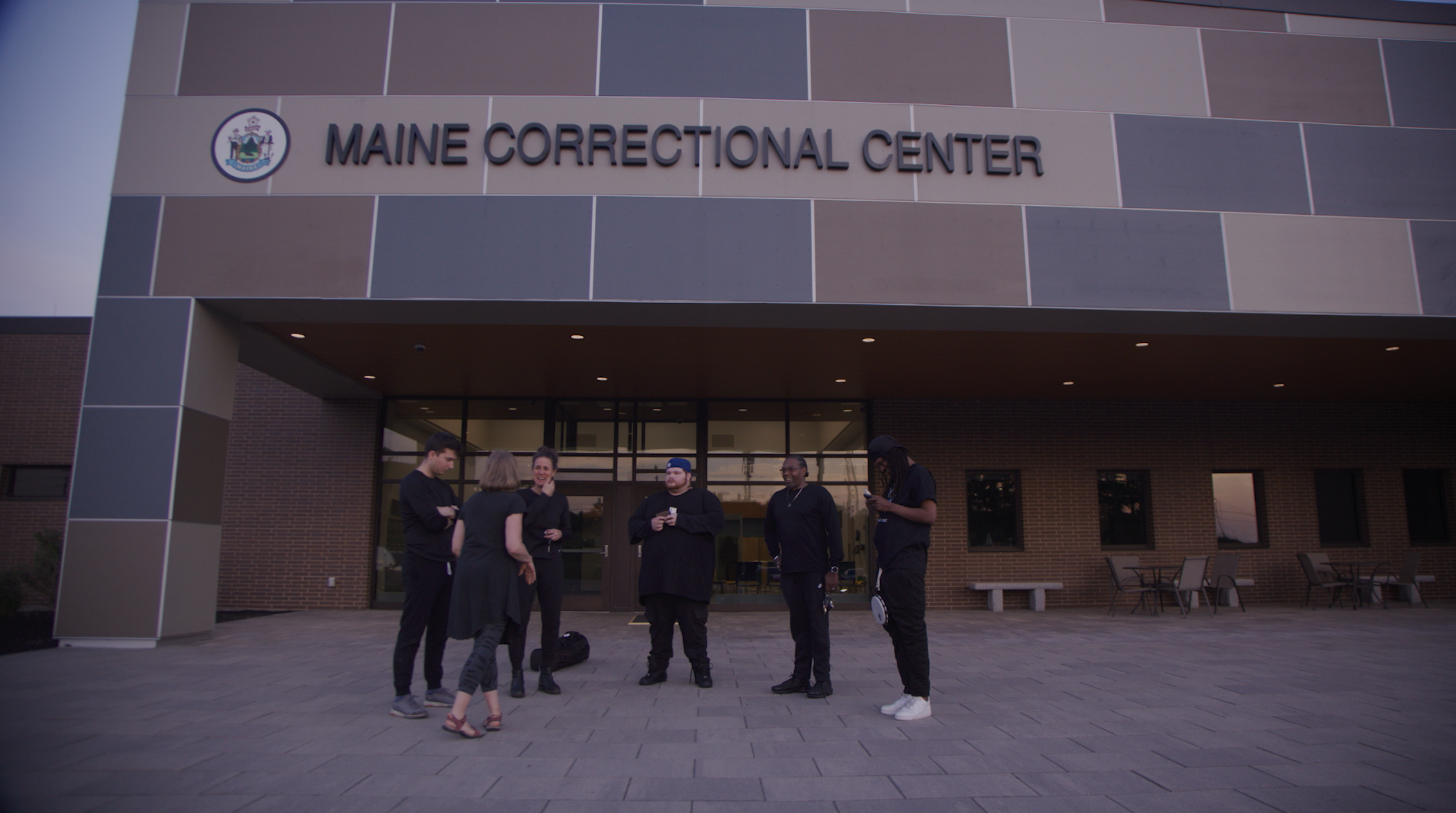 A group of five people standing outside the Maine Correctional Center, engaging in conversation near the building's entrance.