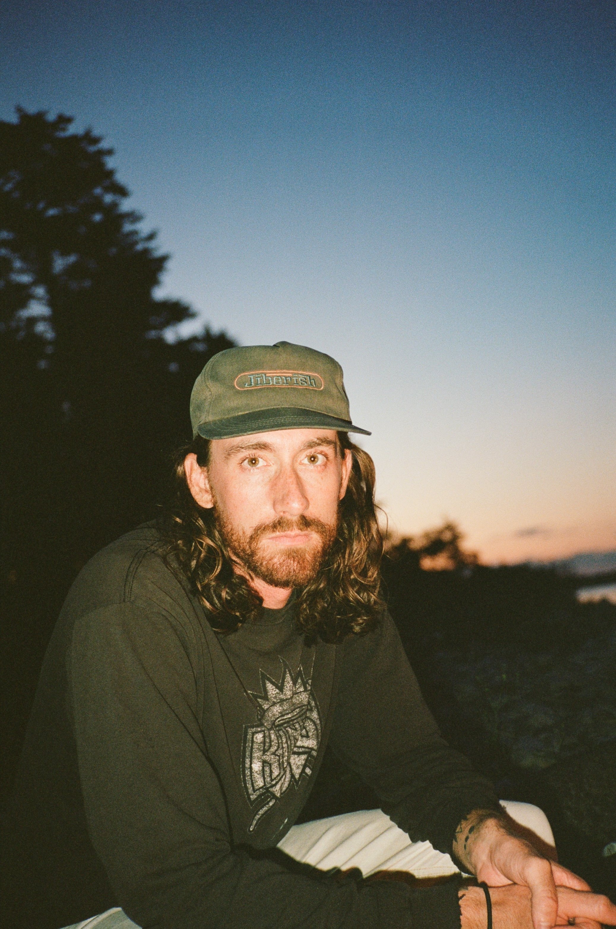 A man with long hair, wearing a green cap and a black shirt with a graphic design, sitting outdoors during sunset with trees in the background.