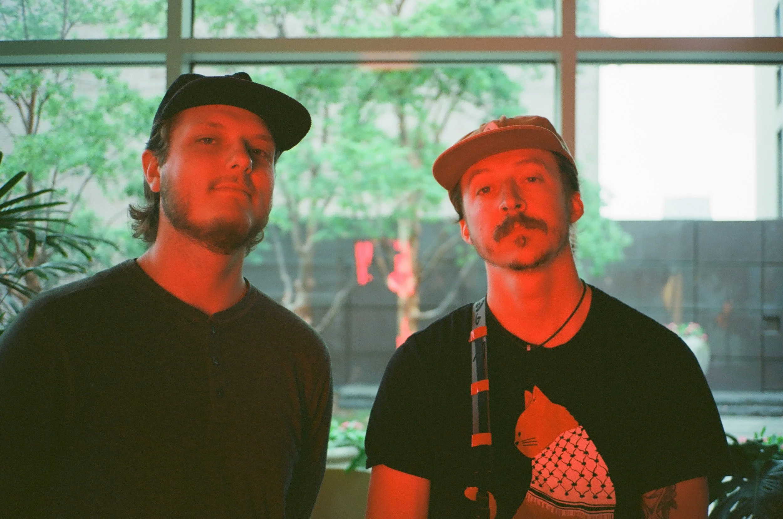 Two young men standing side by side indoors, with large windows and green trees in the background. The man on the left wears a black cap and a dark shirt, while the man on the right wears an orange cap and a black shirt with a graphic design, both looking at the camera.