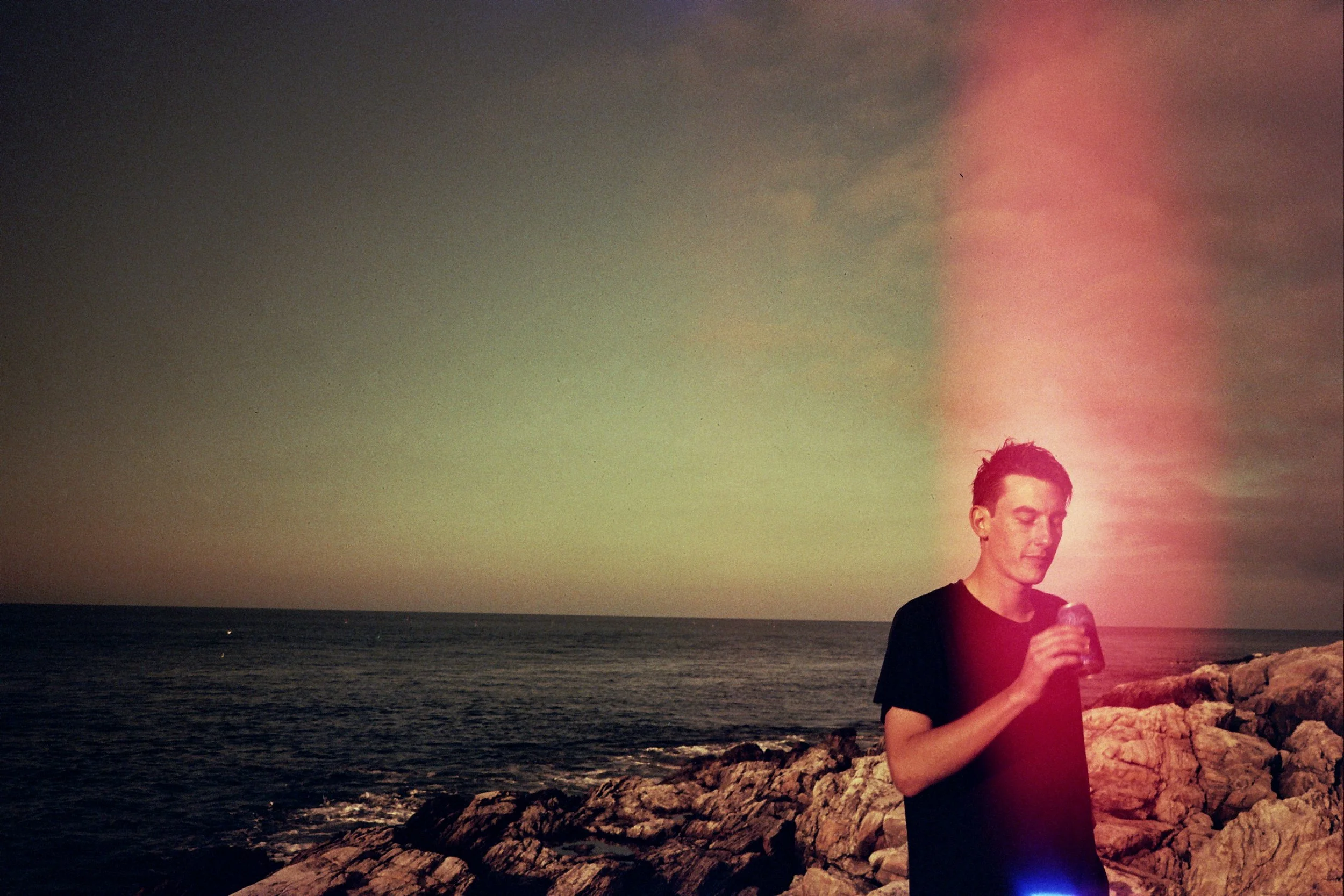 A young man holding a drink, standing on a rocky shoreline at sunset with the ocean in the background.