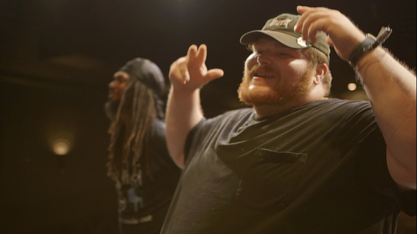 A man wearing a baseball cap, black T-shirt, and wristwatch smiling and raising his hands in the air, with another person with dreadlocks in the background.