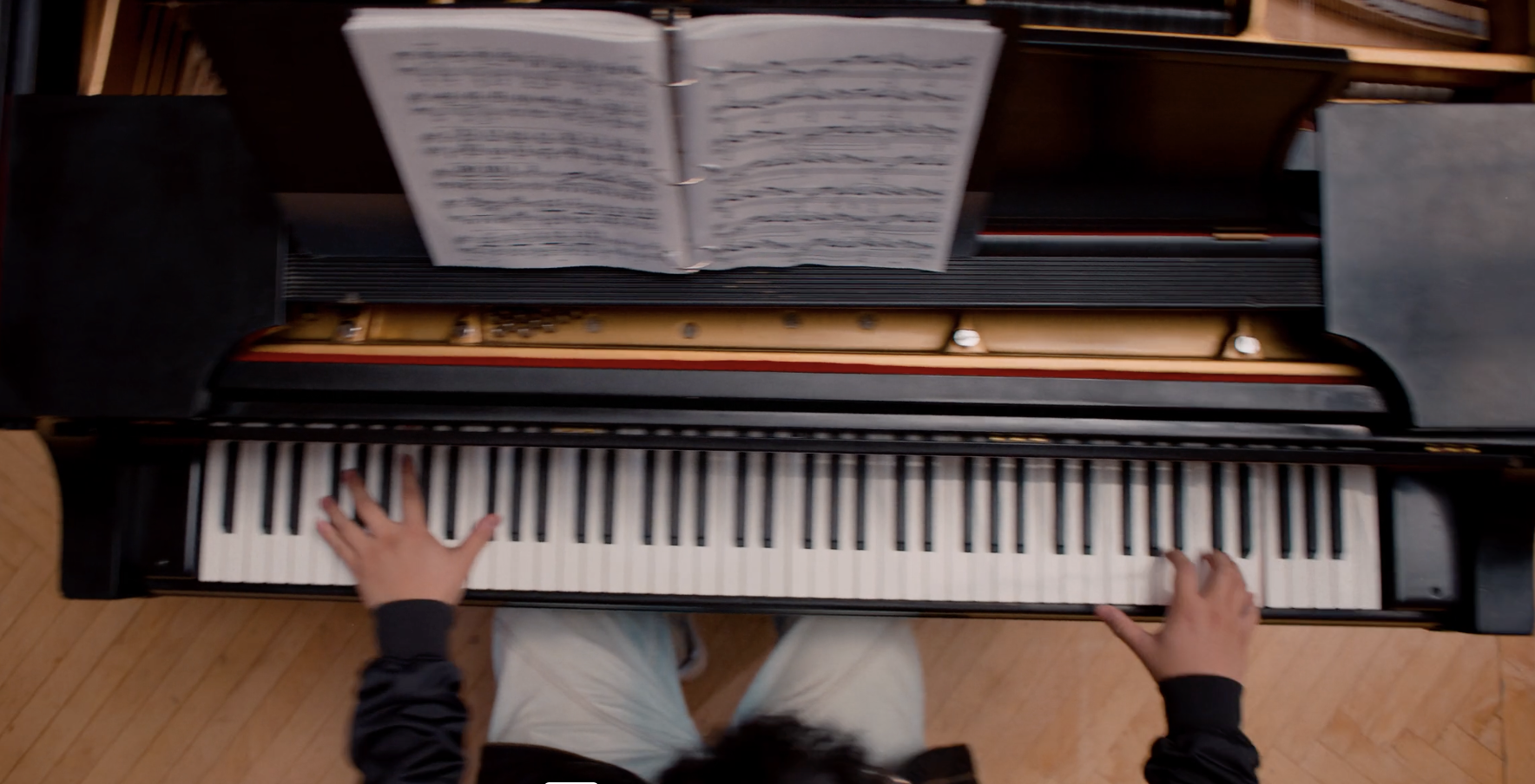 Top view of a person playing a grand piano, with sheet music and a music stand on the piano. The person has their left hand on the keyboard and their right hand is near the middle of the keyboard. The person is wearing a dark long-sleeve shirt and light-colored pants, with a wooden floor underneath.