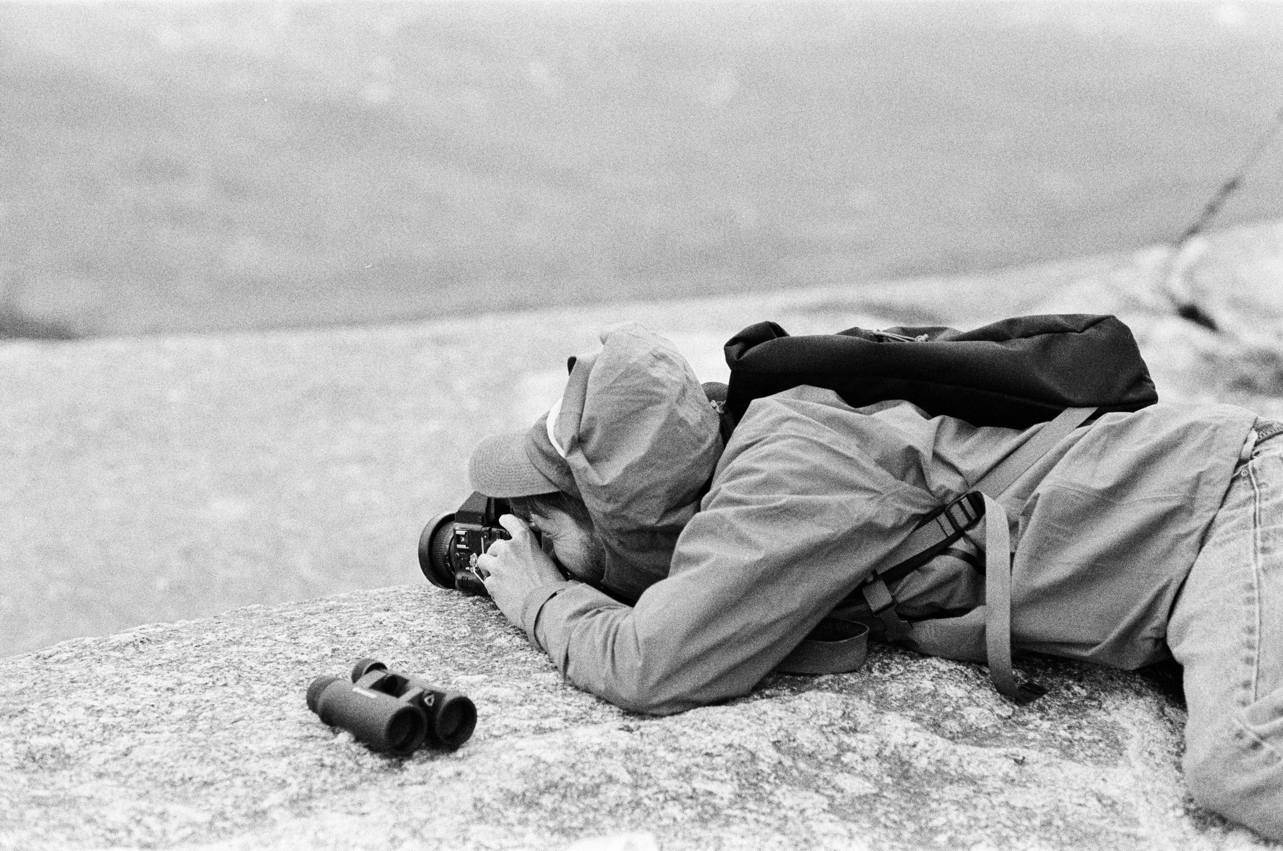 Person lying on the ground outdoors, taking a photograph with a camera, with binoculars on the ground nearby.