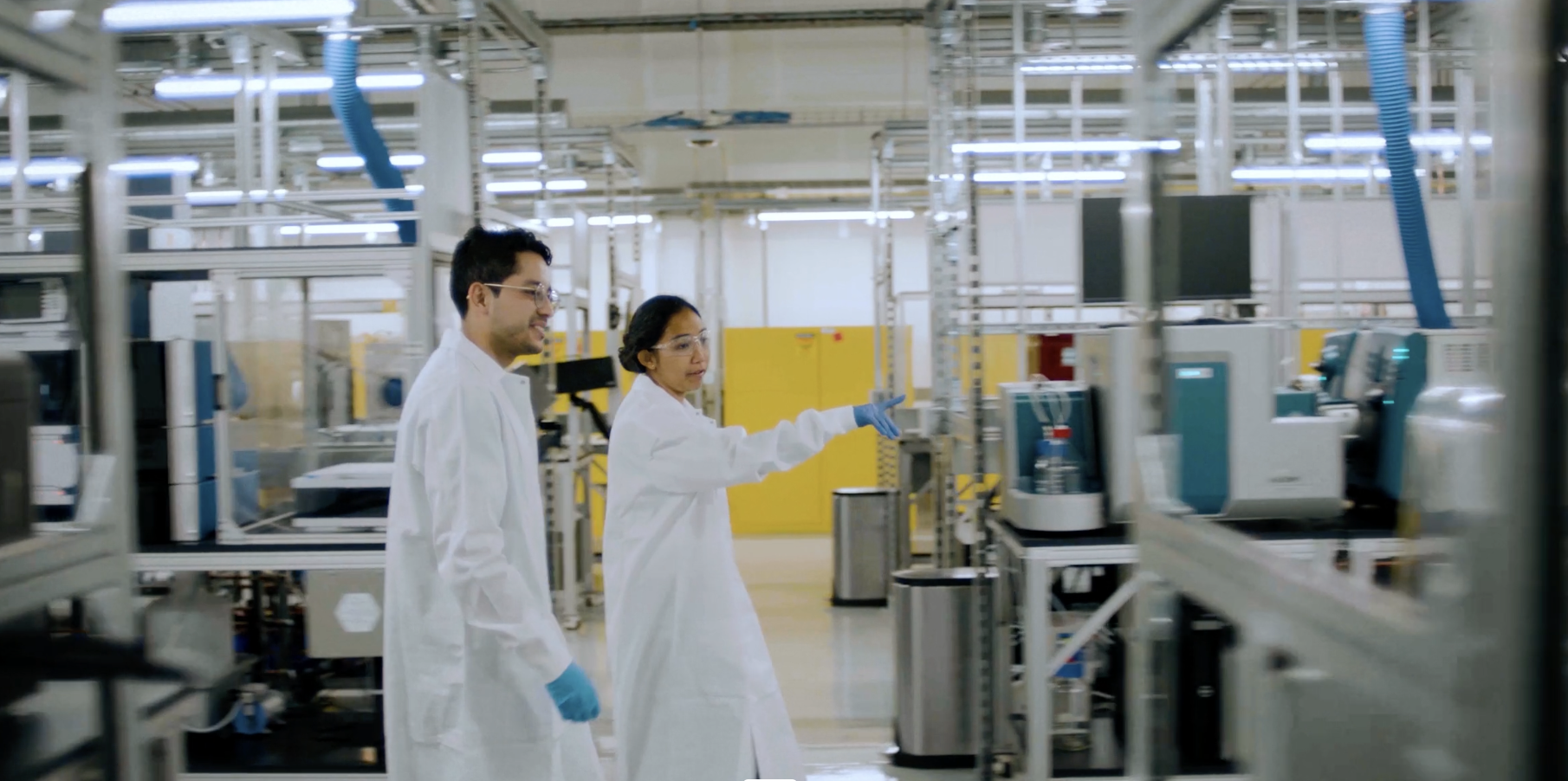 Two scientists, a man and a woman, wearing lab coats, gloves, and safety glasses, working in an industrial laboratory or manufacturing facility with advanced equipment and machinery.