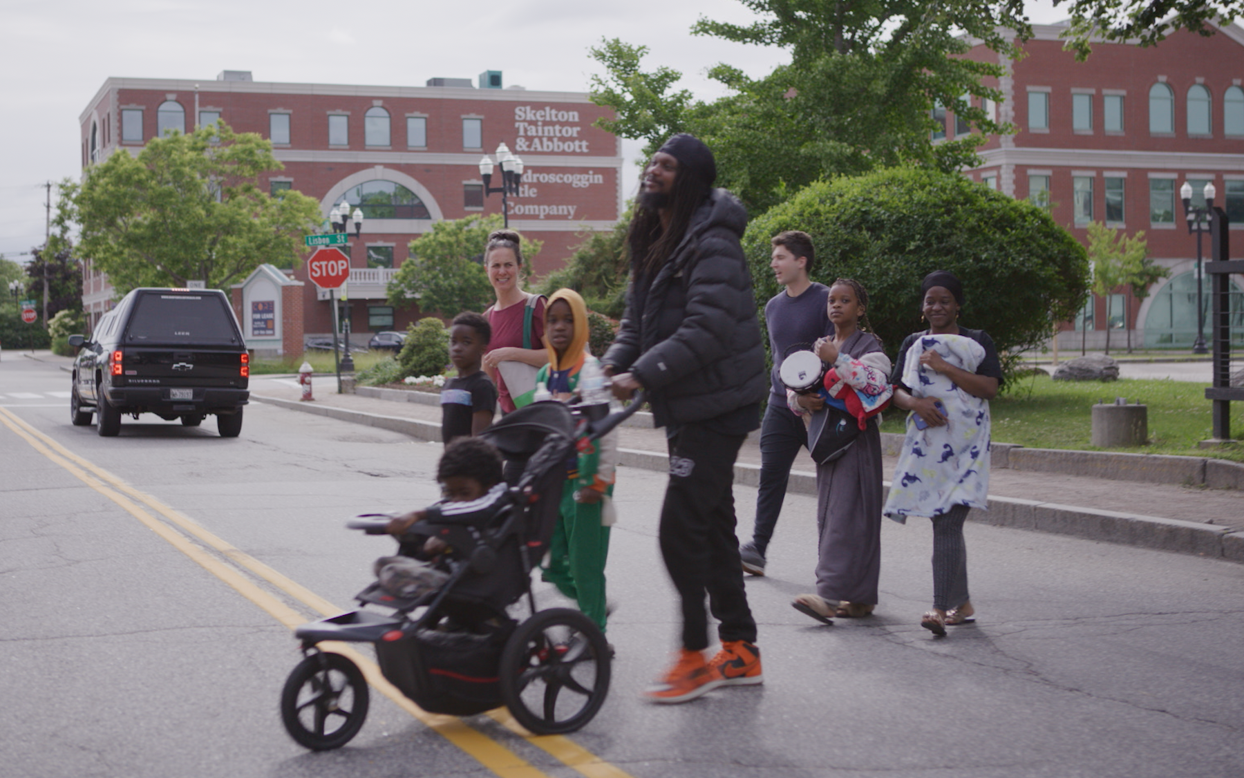 A diverse group of people crossing a street at a crosswalk in an urban area during daytime. Included are children, a man with a stroller, and adults walking together, with buildings and a stop sign in the background.