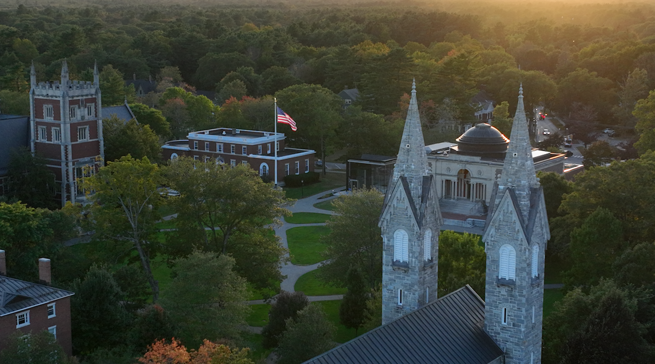 Aerial view of a church with tall stone steeples, surrounded by trees, a building with a flag, and a rural landscape at sunset.