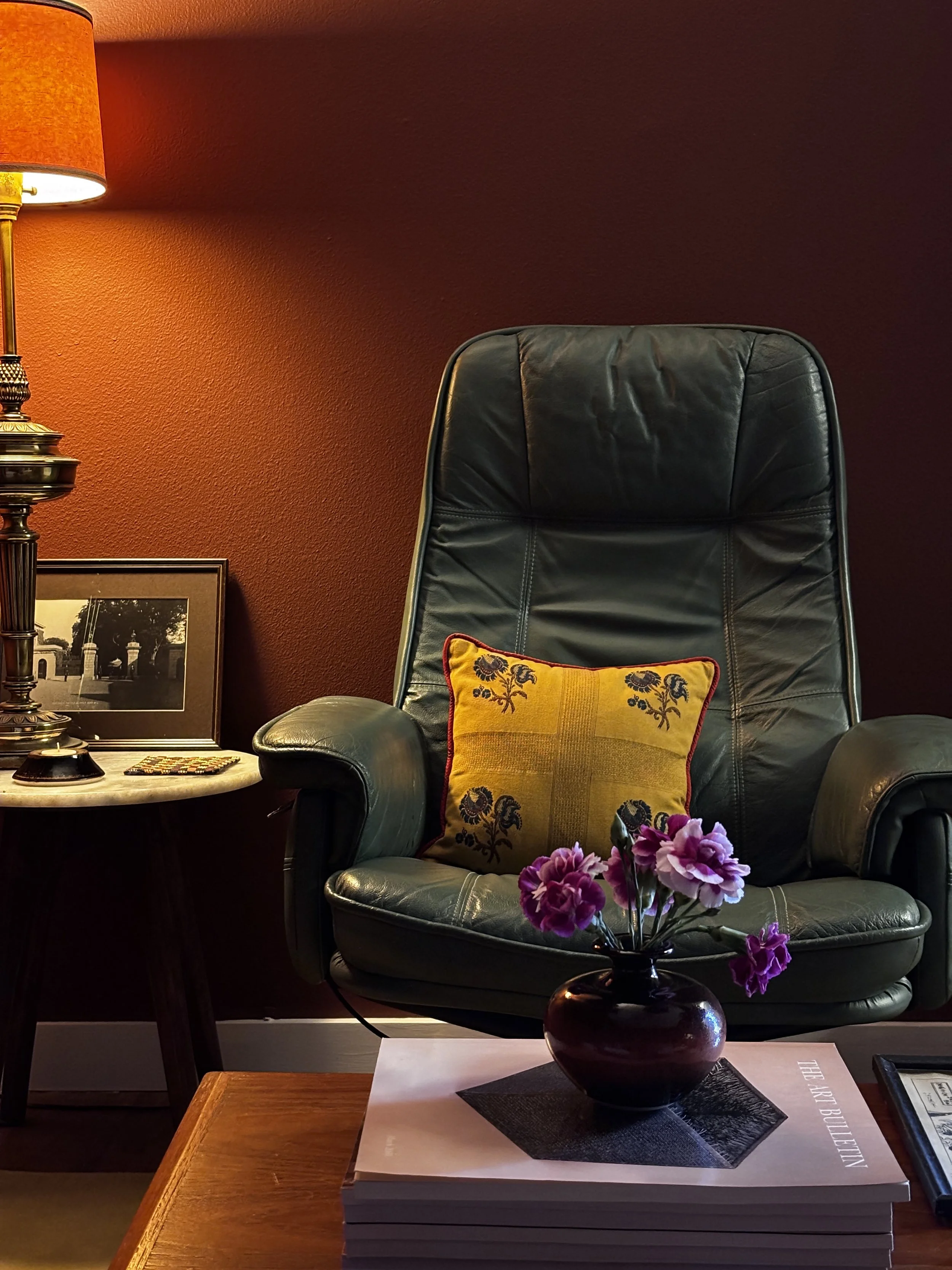 A black leather armchair with a yellow cushion with floral embroidery, a table lamp, a framed picture, and a stack of books with a vase of pink flowers in the foreground, against a dark red wall.