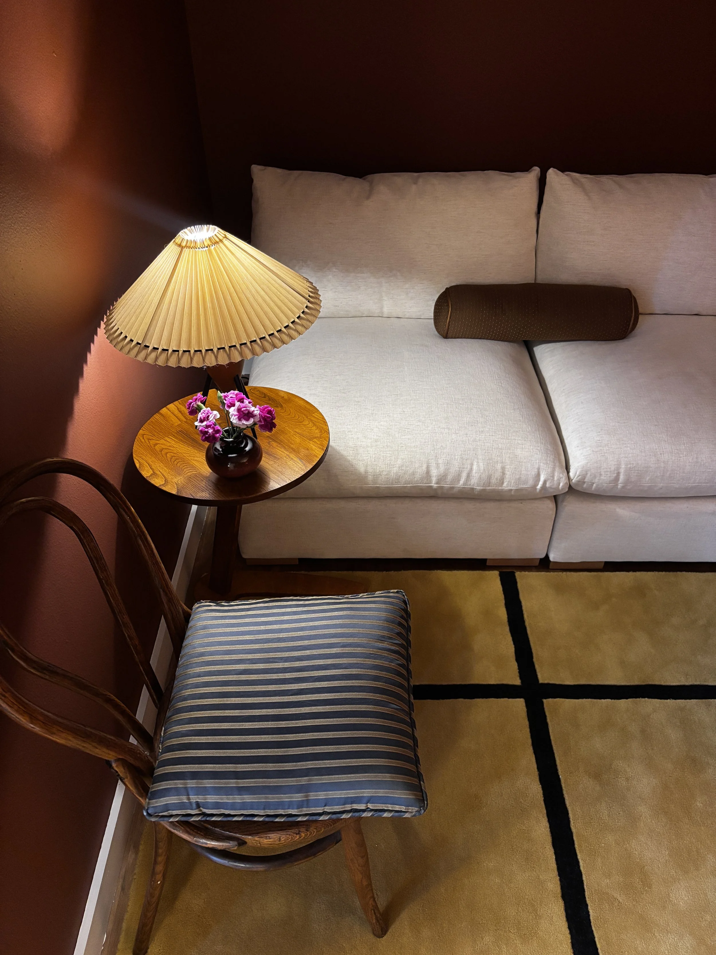A cozy living room corner featuring a woven armchair with a striped cushion, a small wooden side table with a lamp and a vase of pink flowers, a white sofa with a dark cylindrical pillow, a patterned rug, and a dark wall background.