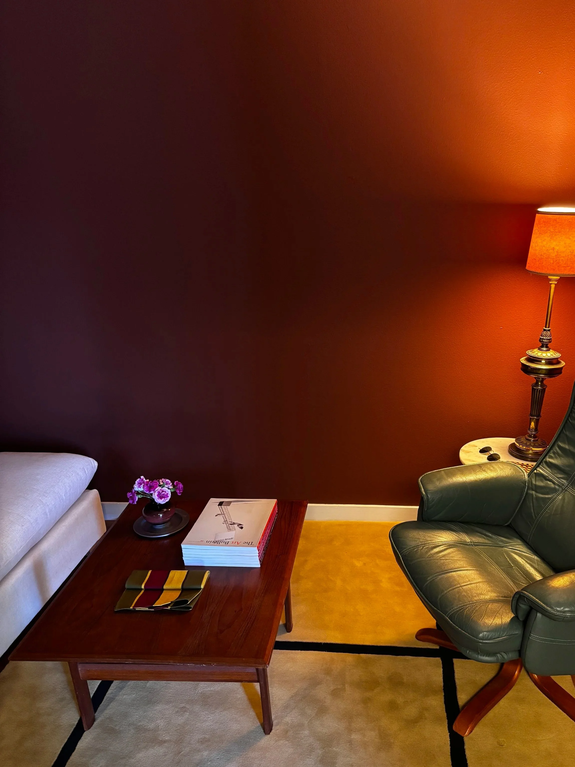 A living room with a dark red wall, a white sofa, a wooden coffee table with magazines and a book, a purple flower vase, a black leather armchair, a side table with a lamp, and a beige carpet.