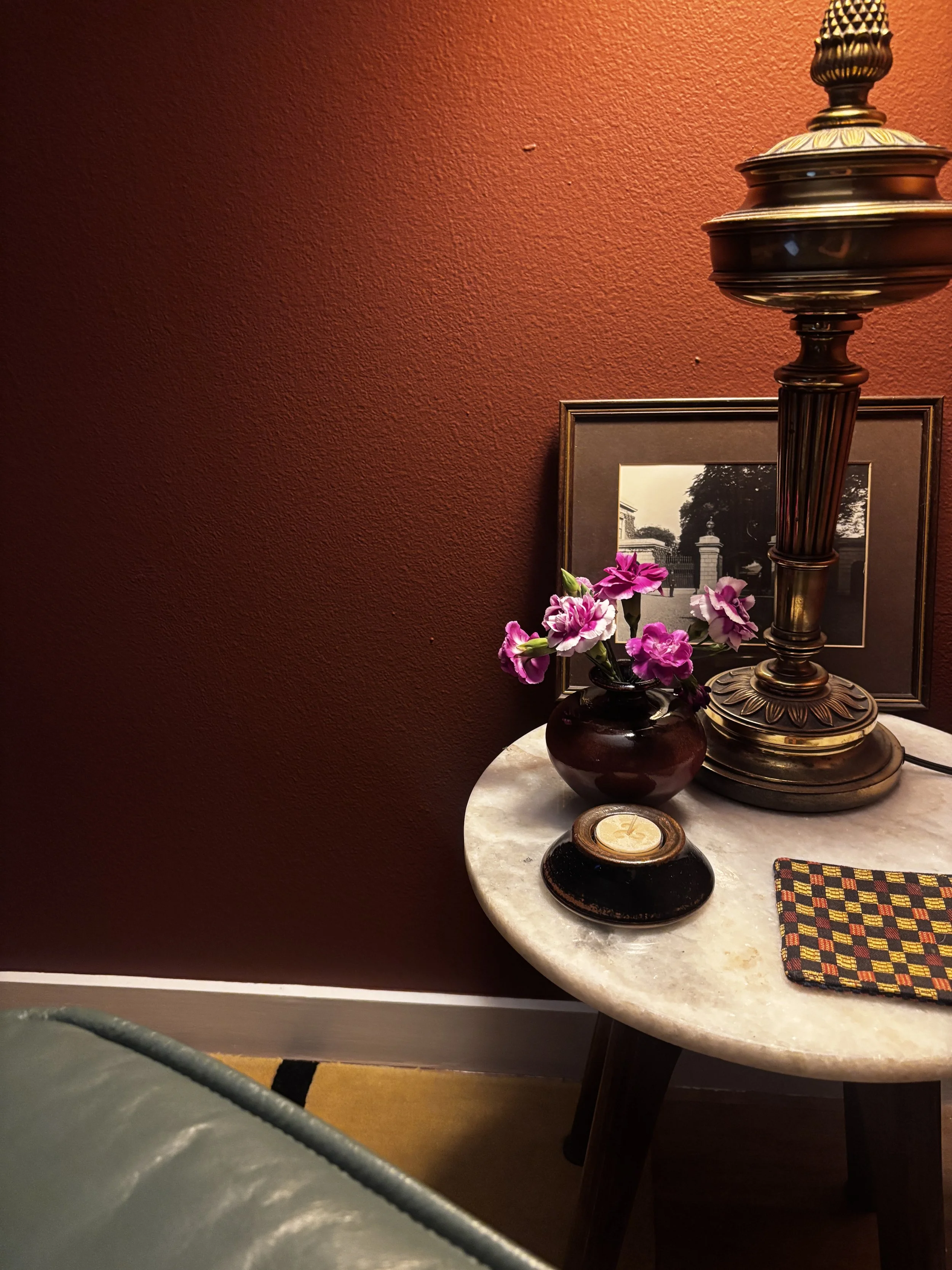 A round marble table with decorative objects including a black vase with pink and white flowers, a small black dish with a candle, a patterned cloth, a framed black-and-white photo of a gate and columns, a tall ornate metal lamp, and a red textured wall in the background.