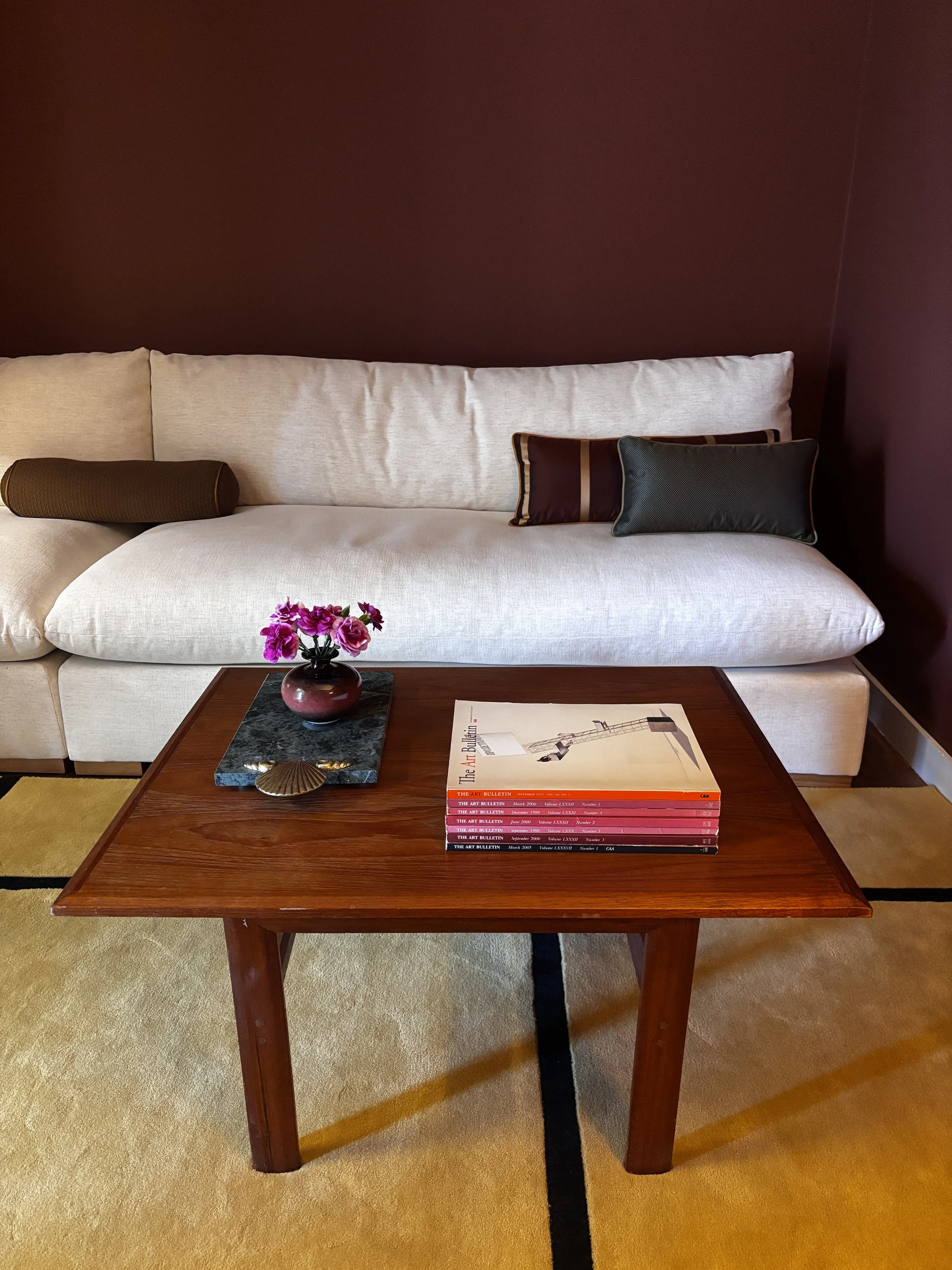 A wooden coffee table with a stack of magazines, a small pink flower arrangement in a dark vase, a shell ornament, and a black-and-beige striped book on top. Behind the table is a white sofa with three large pillows and a cylindrical brown bolster. The sofa is against a dark red wall and the room has a beige carpet.