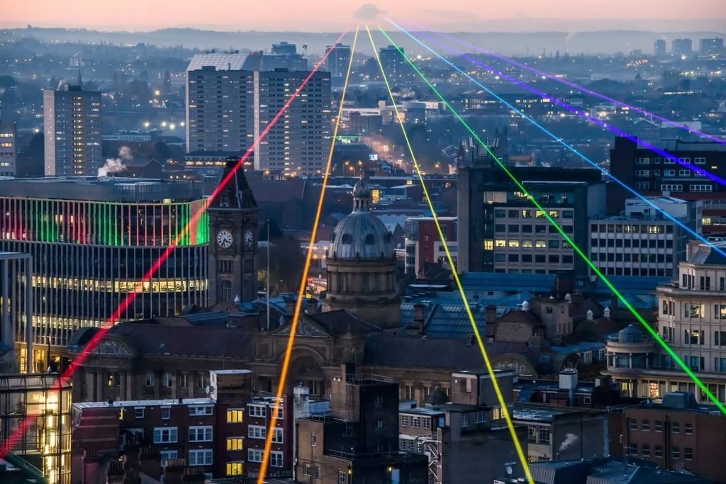 Birmingham City skyline at dusk with colorful laser light beams crossing the sky and pointing towards various buildings.