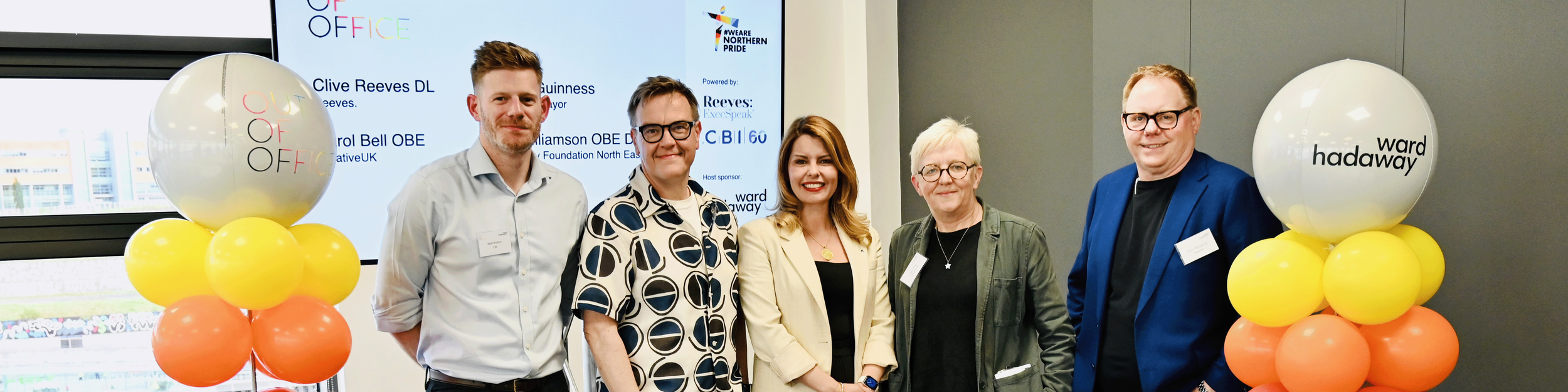 Group of five people standing indoors, smiling for a photo, with large colorful balloons and a presentation screen behind them at an event.