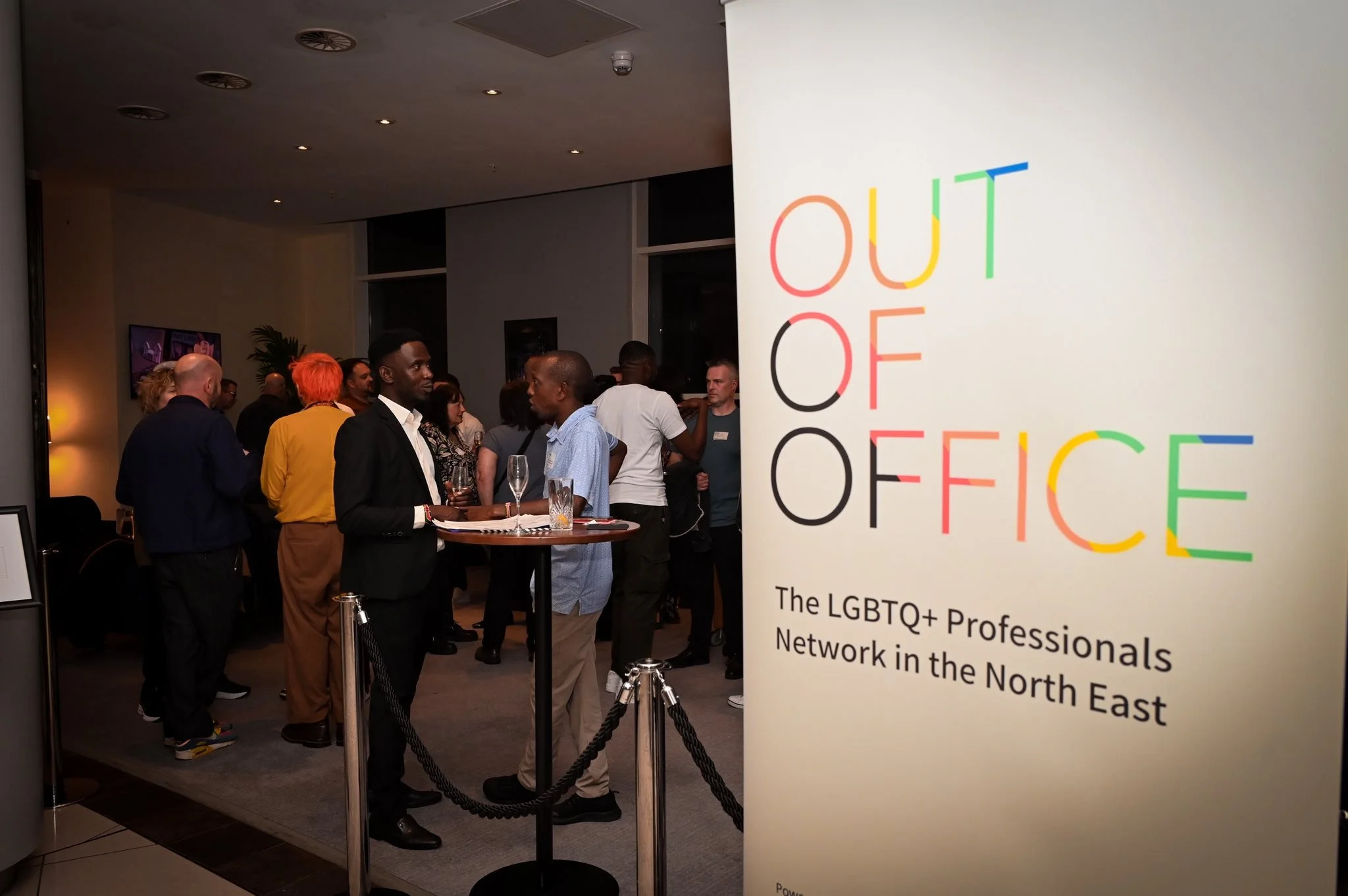 A diverse group of LGBTQ+ professionals at a networking event in a modern indoor setting, with a prominent sign reading 'OUT OF OFFICE' and describing the LGBTQ+ Professionals Network in the North East.