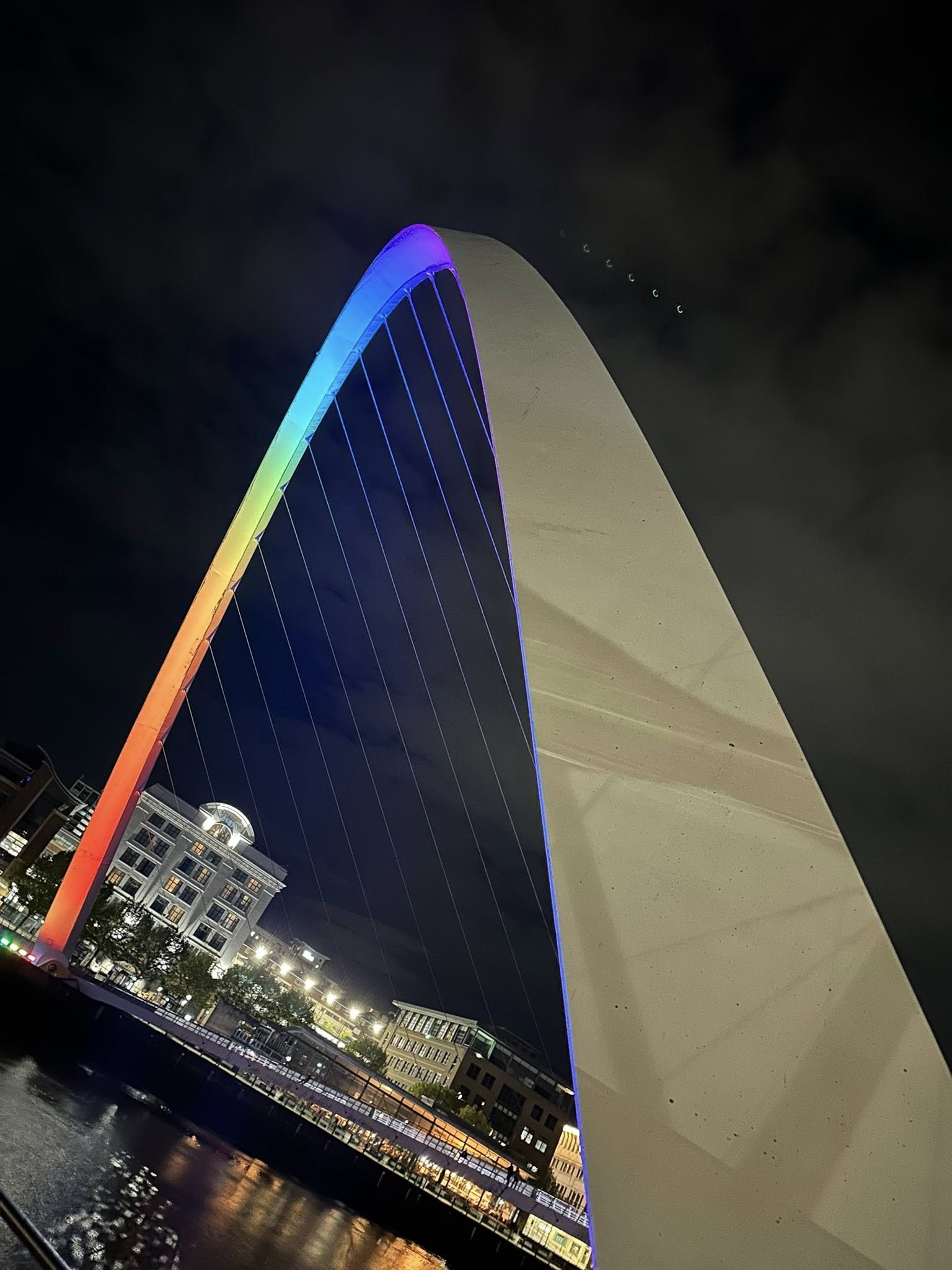 Night view of the Gateshead Millennium Bridge, illuminated with rainbow-colored lights, reflected in the River Tyne.