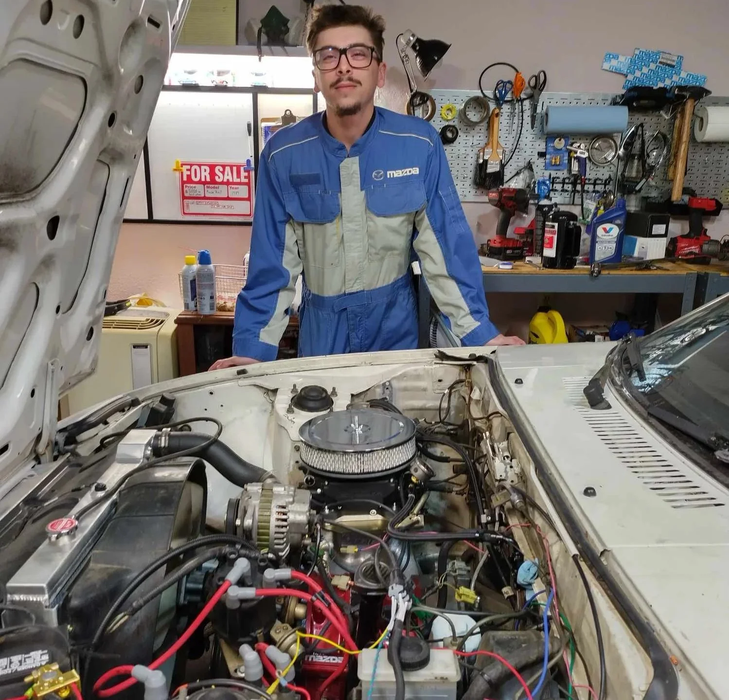 A man in a blue racing suit leaning over an open car hood in a garage, with tools and equipment on a workbench behind him.