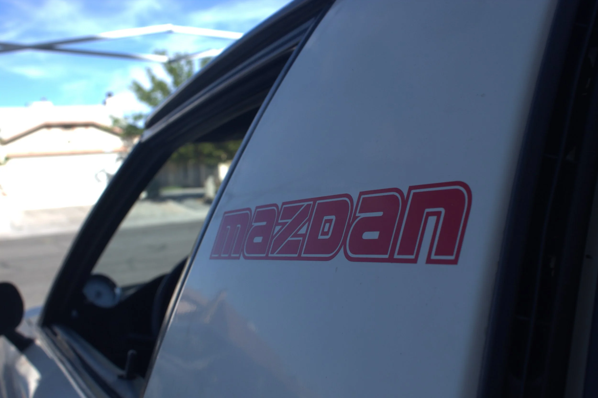 Close-up of a Mazda vehicle with the Mazda logo visible on the side, slightly tilted, with a background of a building and blue sky.