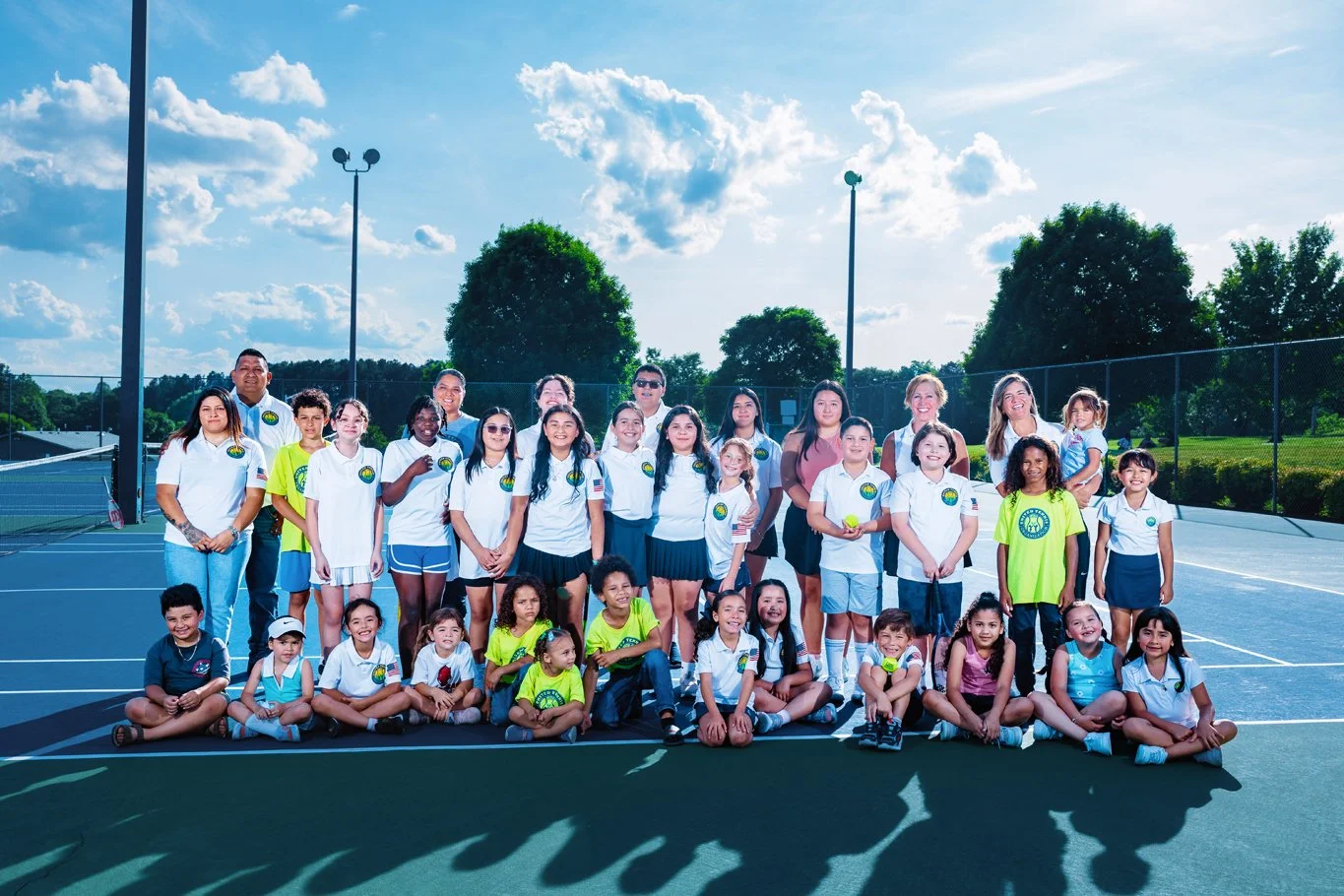 Group photo of children and adults on a tennis court, some catching tennis balls, outdoors on a sunny day with trees and a blue sky with clouds in the background.