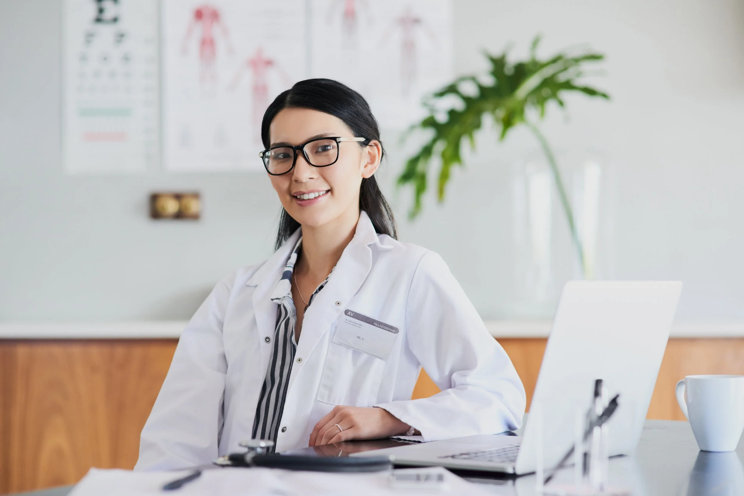 Smiling female doctor or healthcare professional with glasses, sitting at a desk with a laptop and medical papers, in a medical office with a herbal poster and large plant in the background.