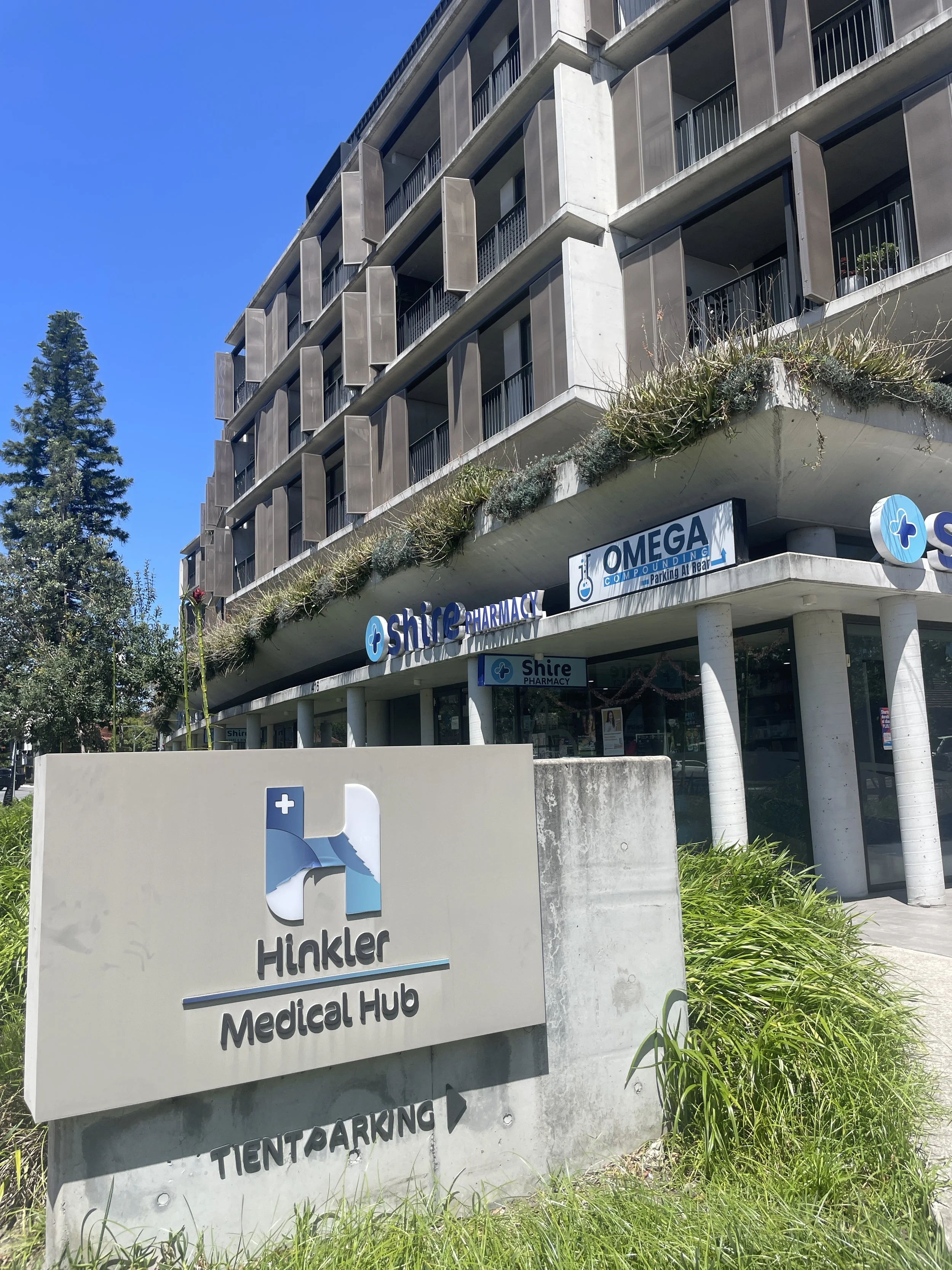 Office building with signage for Hikmler Medical Hub, Shire Pharmacy, and Omega Compounding, with a parking nearby, under a clear blue sky.