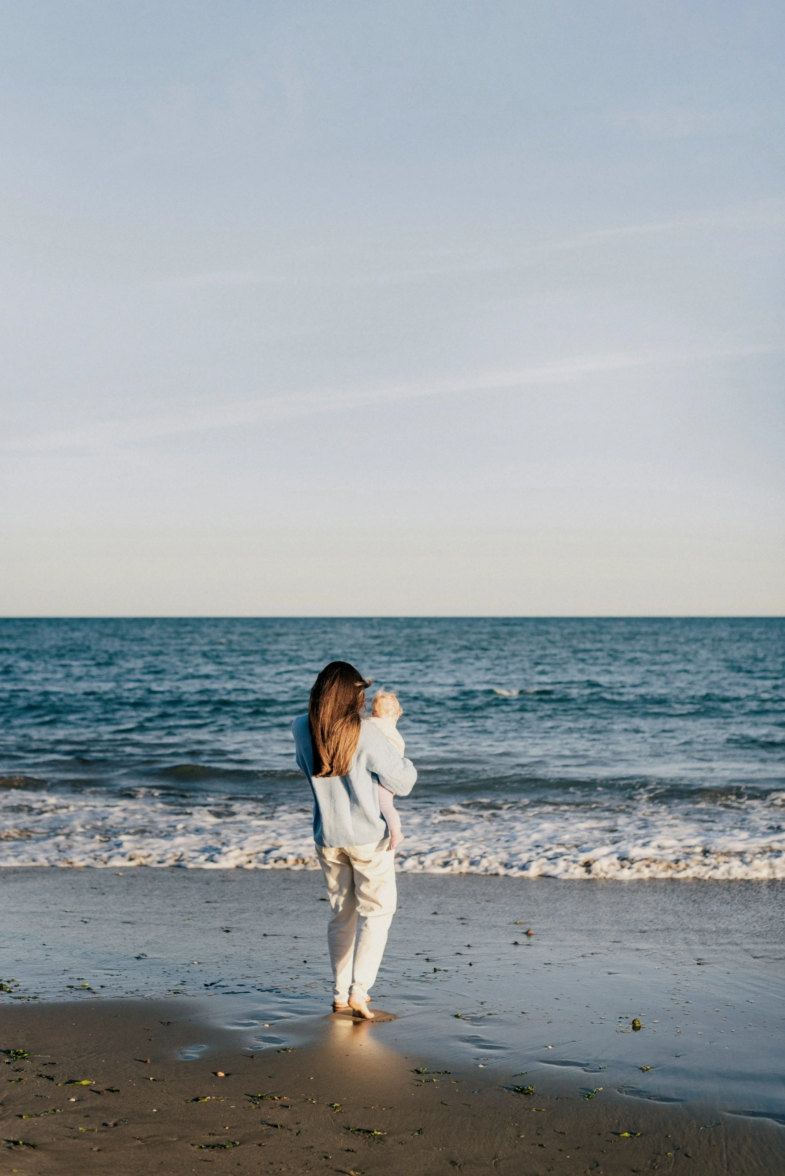 A woman holding a child by the beach at sunset.