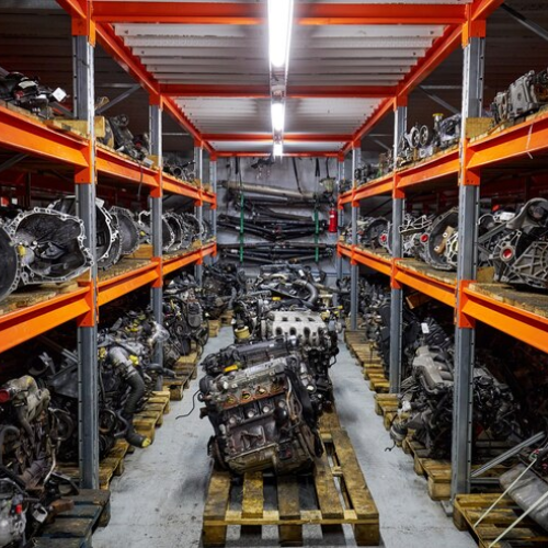 A storage area with automotive engines on orange steel shelves, with many engines on wooden pallets on the floor between shelves.