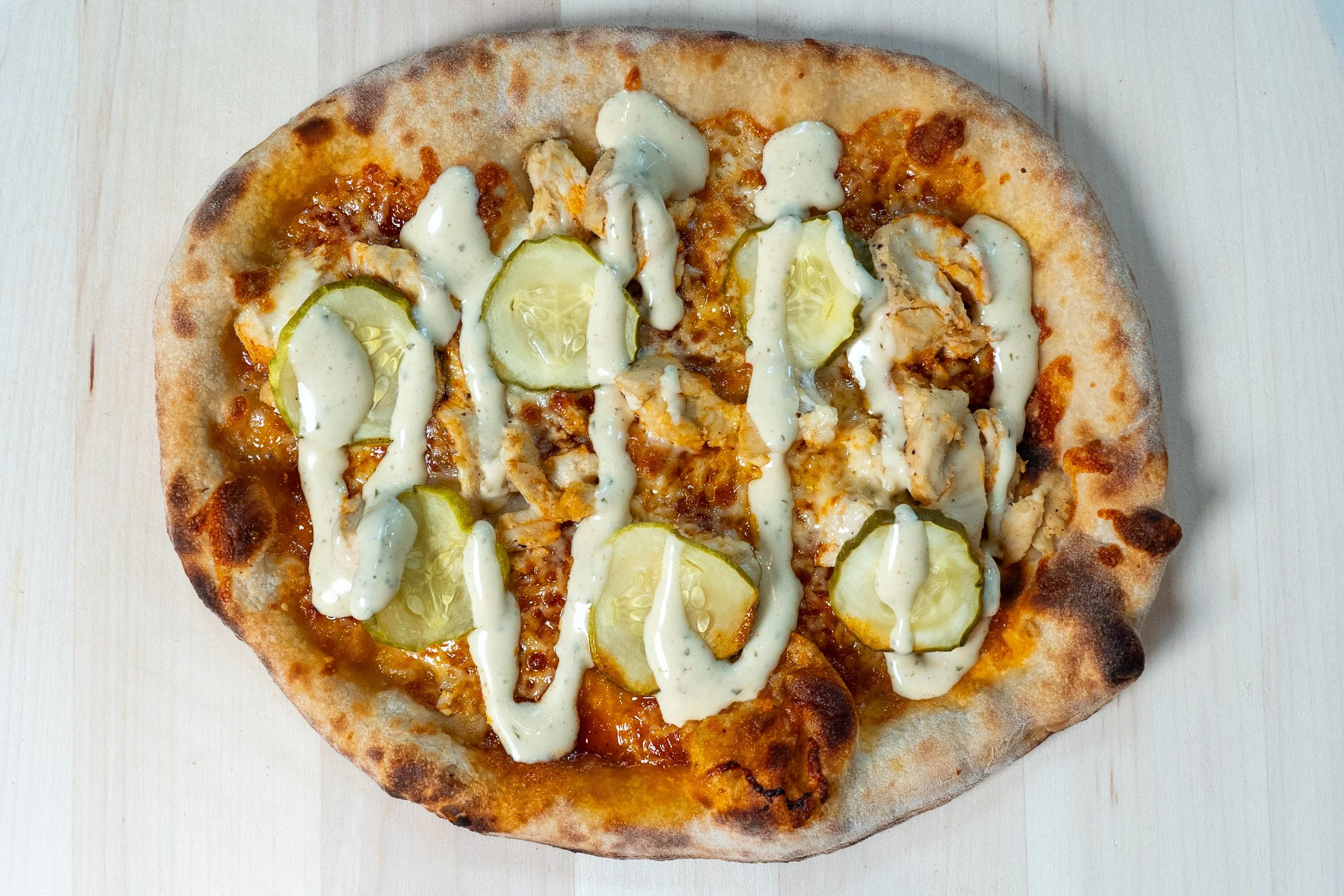 Pizza with sliced pickles, chicken pieces, cheese, and drizzled sauce on top.