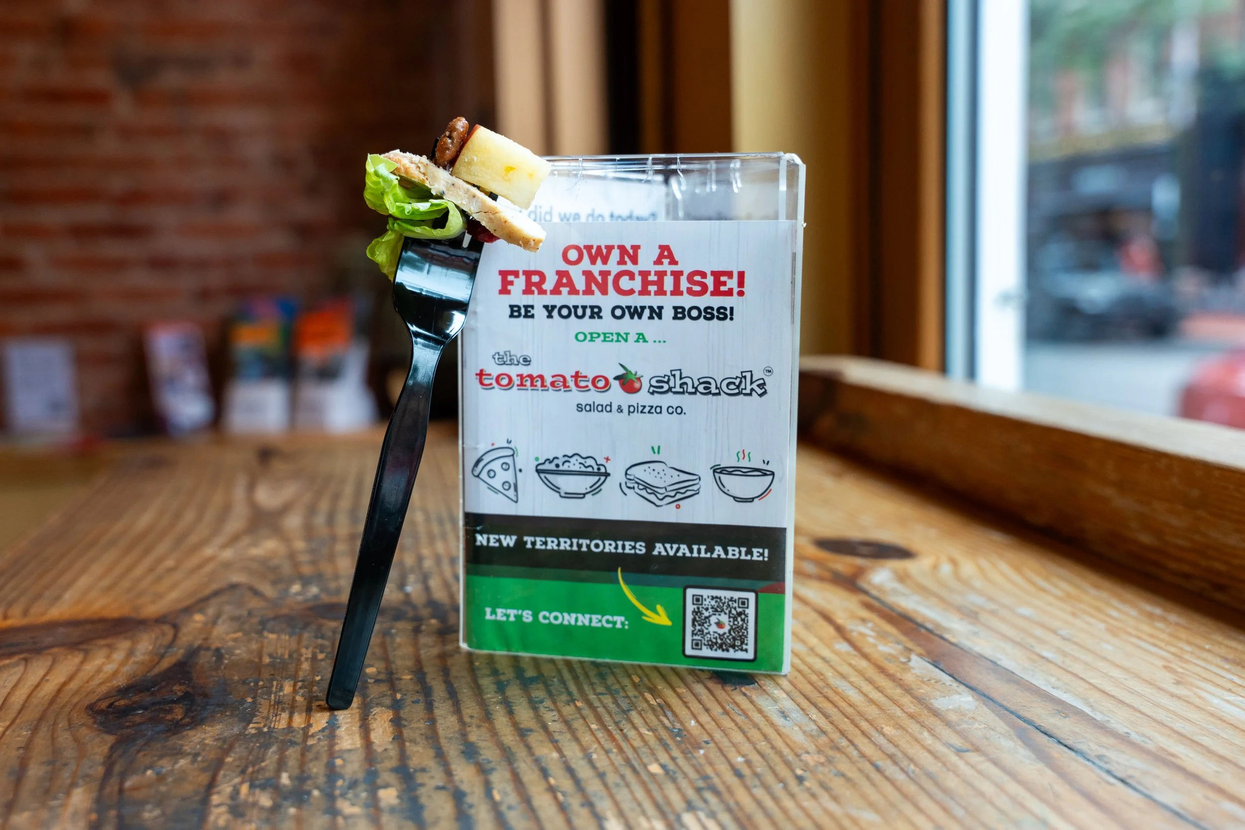 Fork with a sandwich on it placed next to a promotional card for The Tomato Shack restaurant, featuring franchise information and a QR code, on a wooden table near a window.