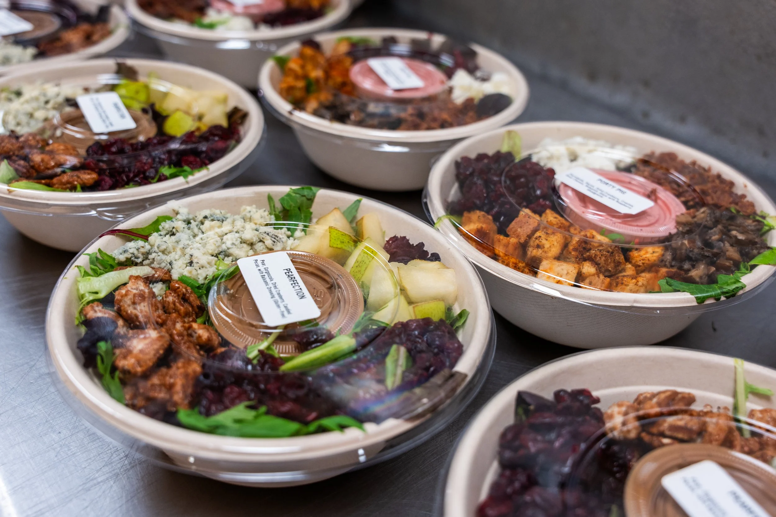 Multiple takeout bowls filled with salads and protein, topped with plastic lids, placed on a metal surface.