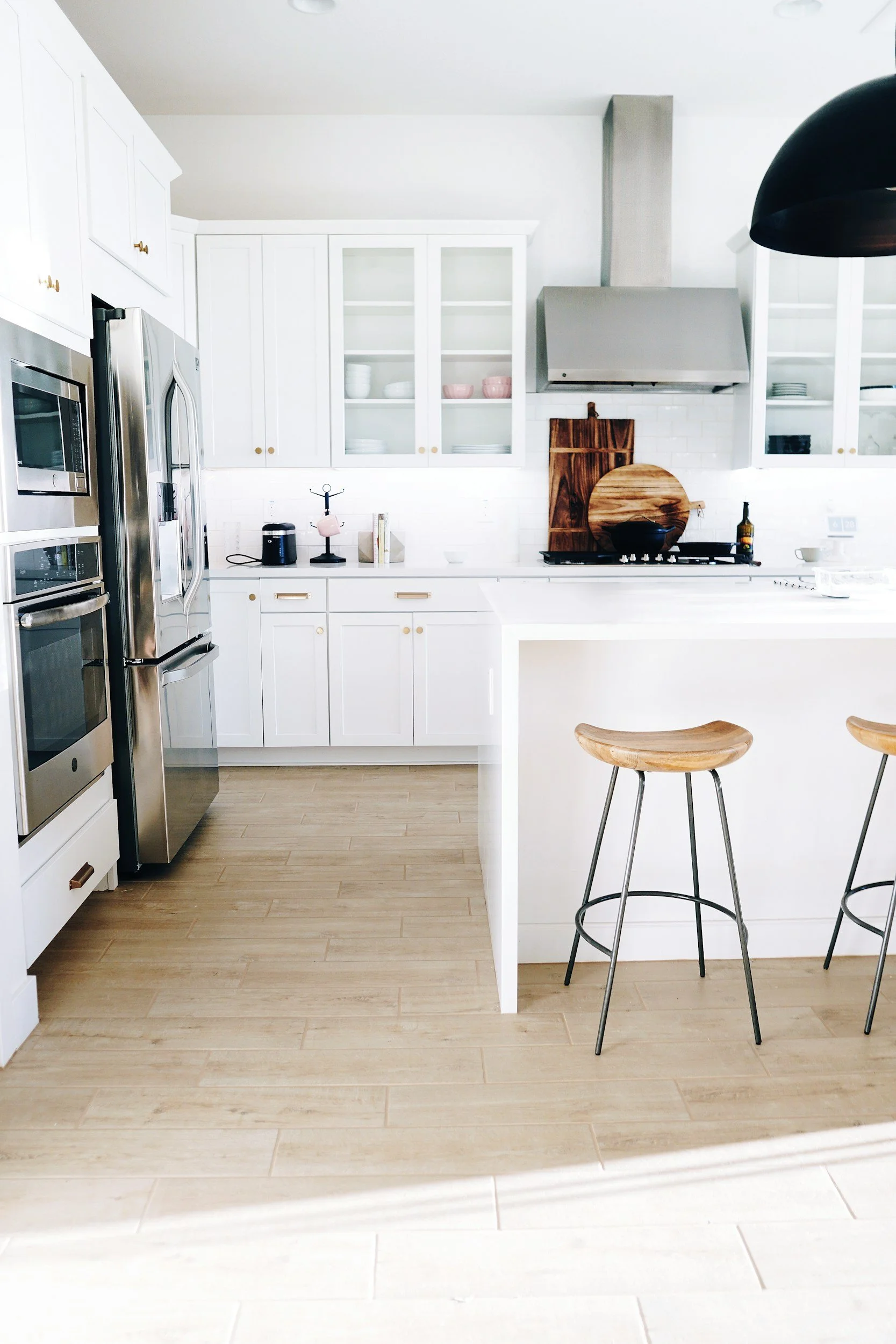 Bright white kitchen with light wood flooring, white cabinets, stainless steel appliances, a kitchen island with wooden bar stools, and a stovetop with a large wooden cutting board and black cookware on it.