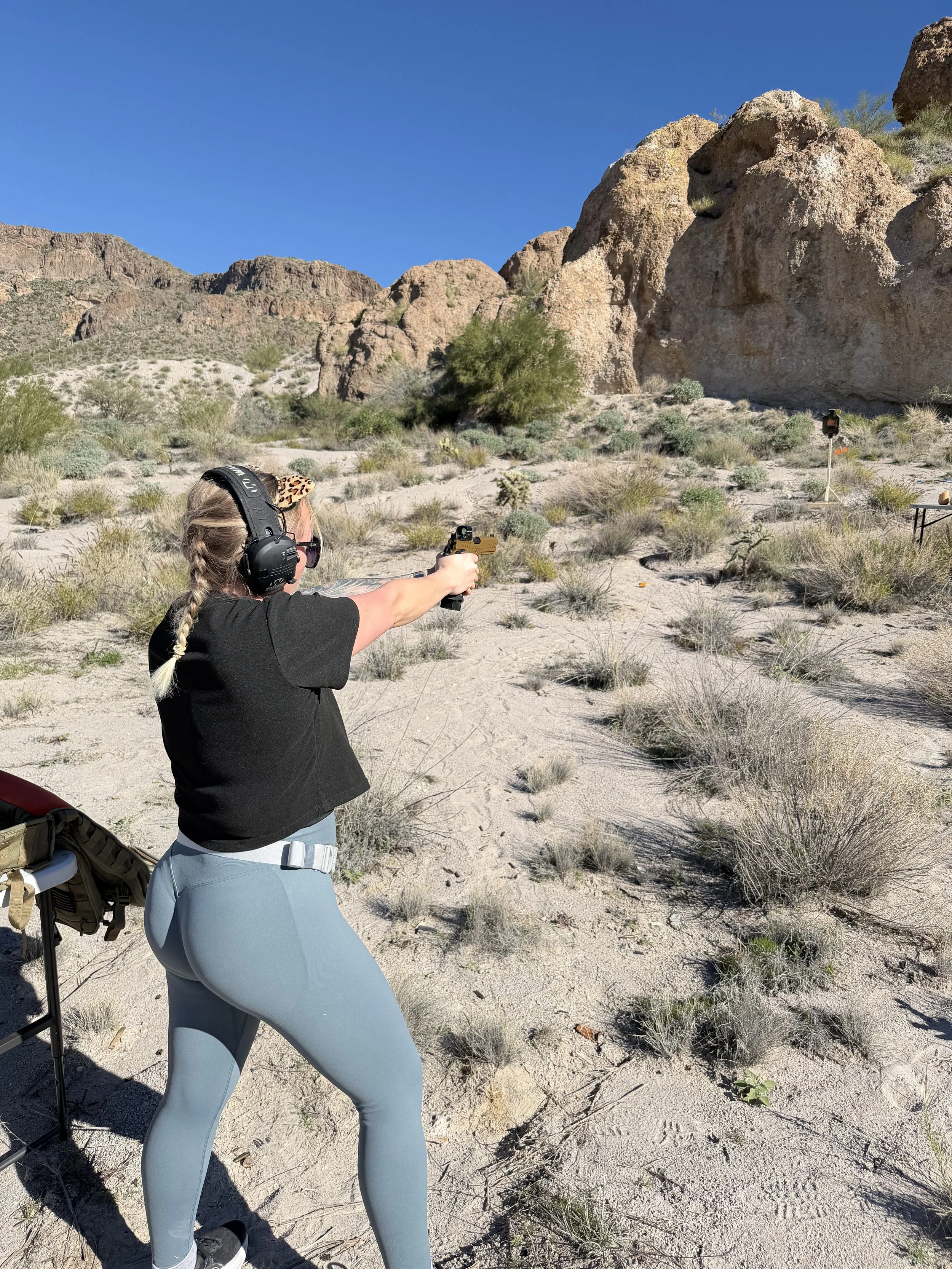 Woman in black t-shirt and light blue leggings shooting a handgun at an outdoor desert shooting range with rocky formations in the background.